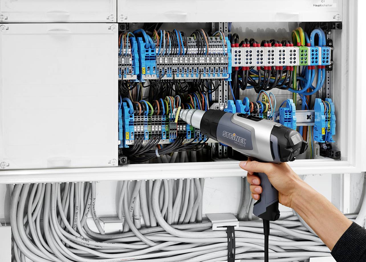 A person is using a heat gun to work on an electrical cabinet with numerous cables and connections.