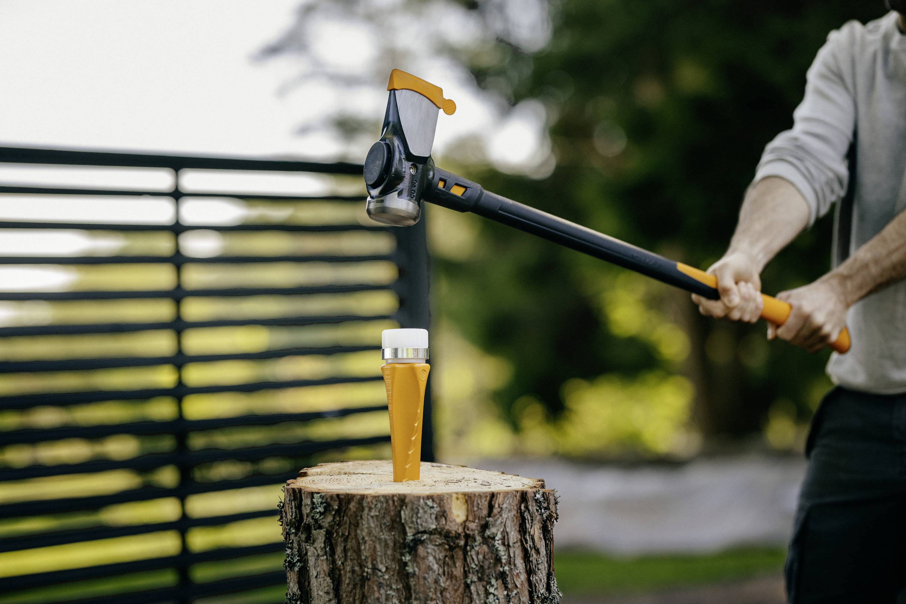 A person is chopping at a tree stump with an axe, with a splitting wedge in place, to split wood, outdoors in daylight.