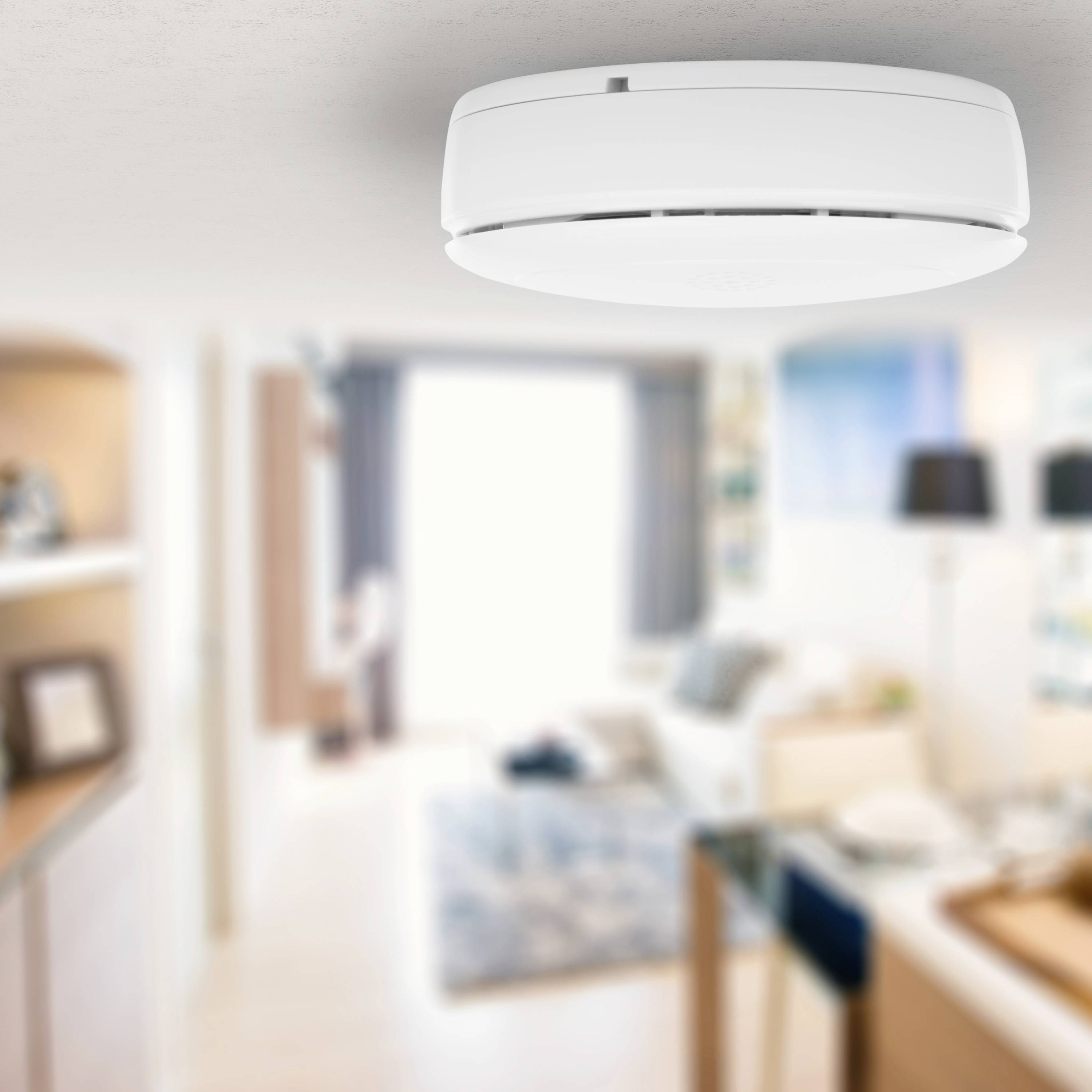 A smoke detector is mounted on the ceiling of a modern living room. The room appears bright and inviting with a sofa and a table.