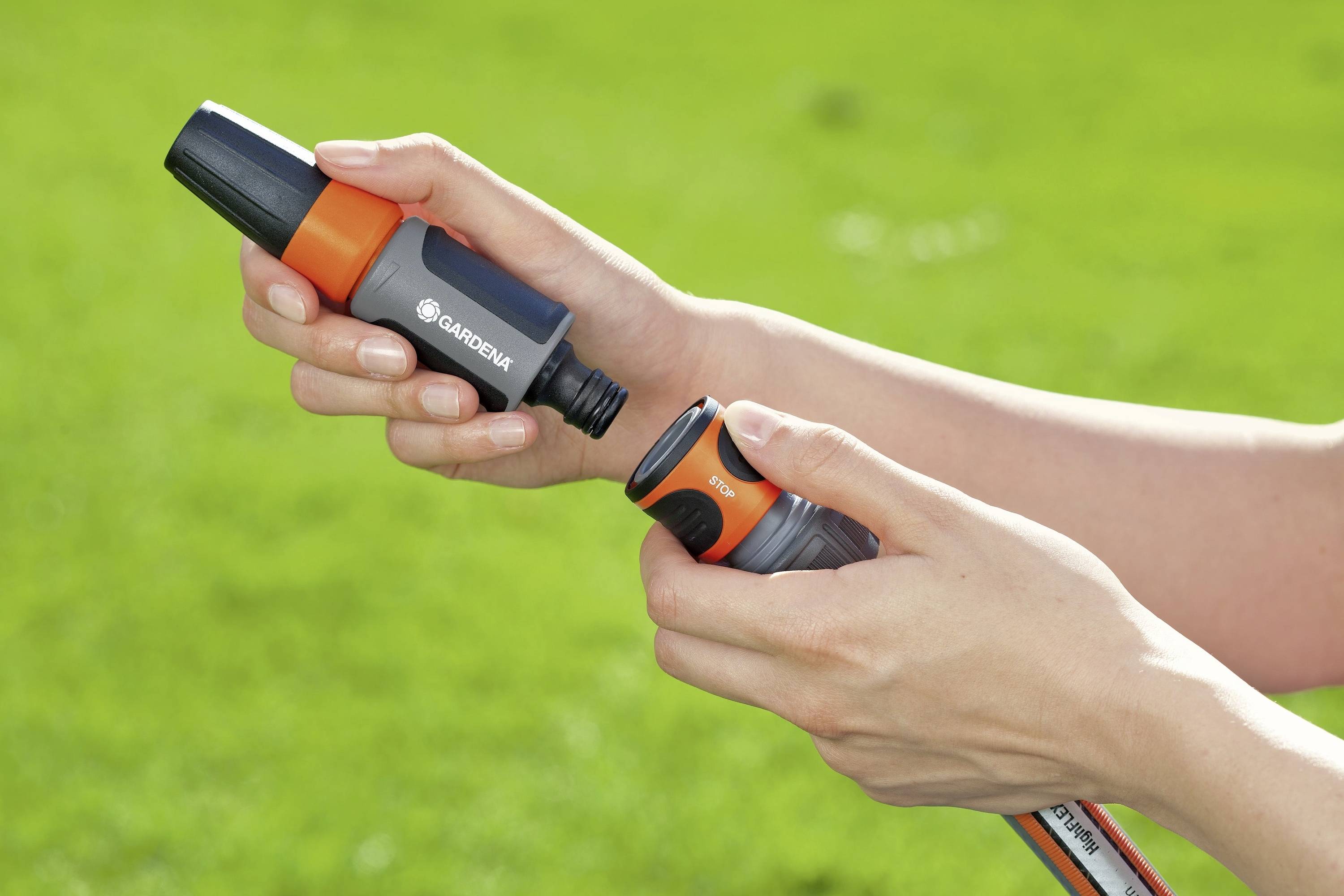 A hand holds a garden hose nozzle, while the other hand grips a connector. The background shows green lawn.