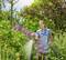 A woman in dungarees waters colourful flowers with a garden hose in a sunny garden full of green plants.