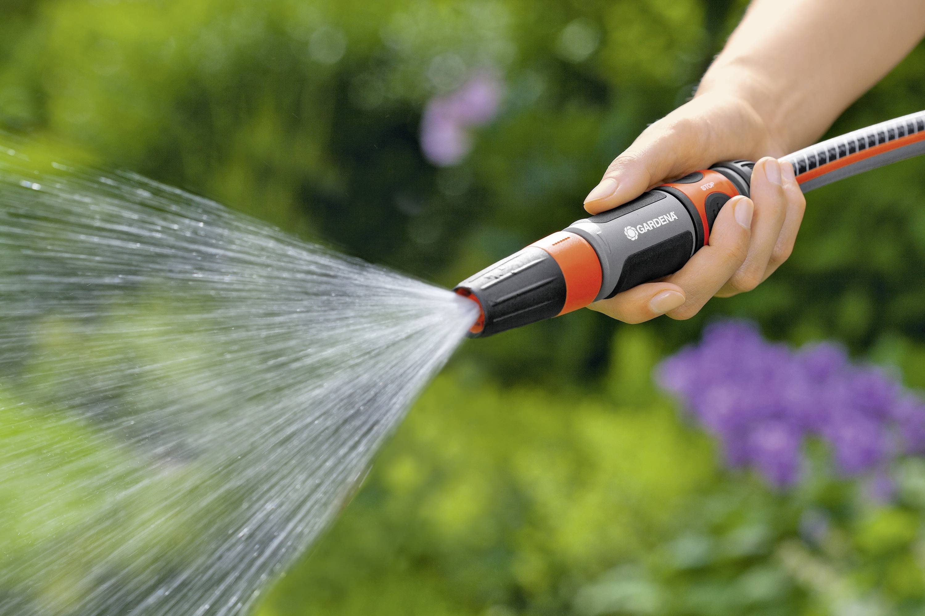 A hand is holding a garden hose, with water spraying from it. Blurred plants are visible in the background, representing garden irrigation.