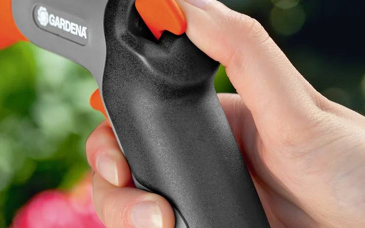 Hand holding a black and orange garden hose nozzle, with a blurred background of greenery and flowers.