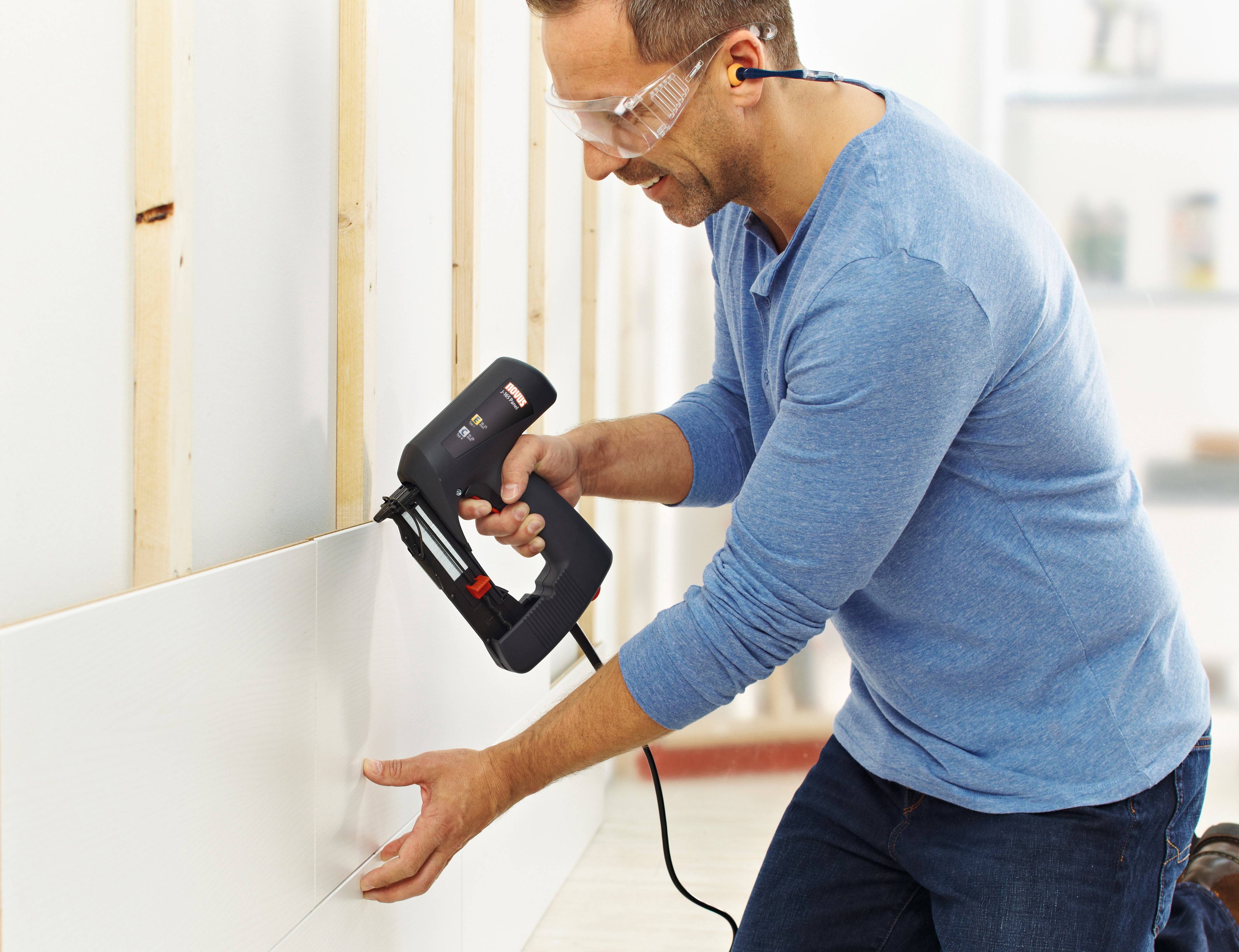A man is using an electric nail gun to secure a board to an open wall frame. He is wearing safety glasses and ear plugs.