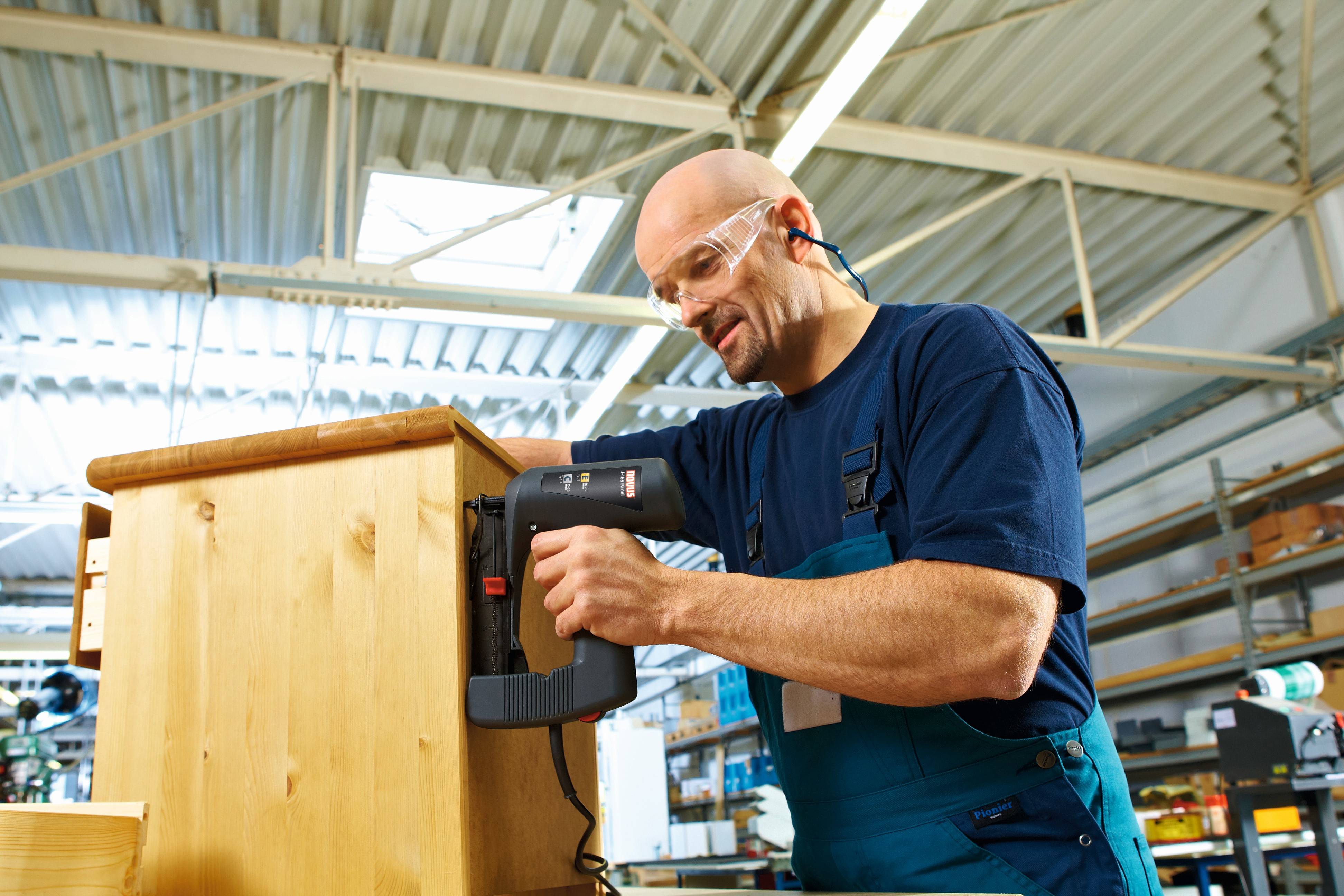 A man in workwear is using an electric tool to assemble a piece of furniture in a bright workshop.