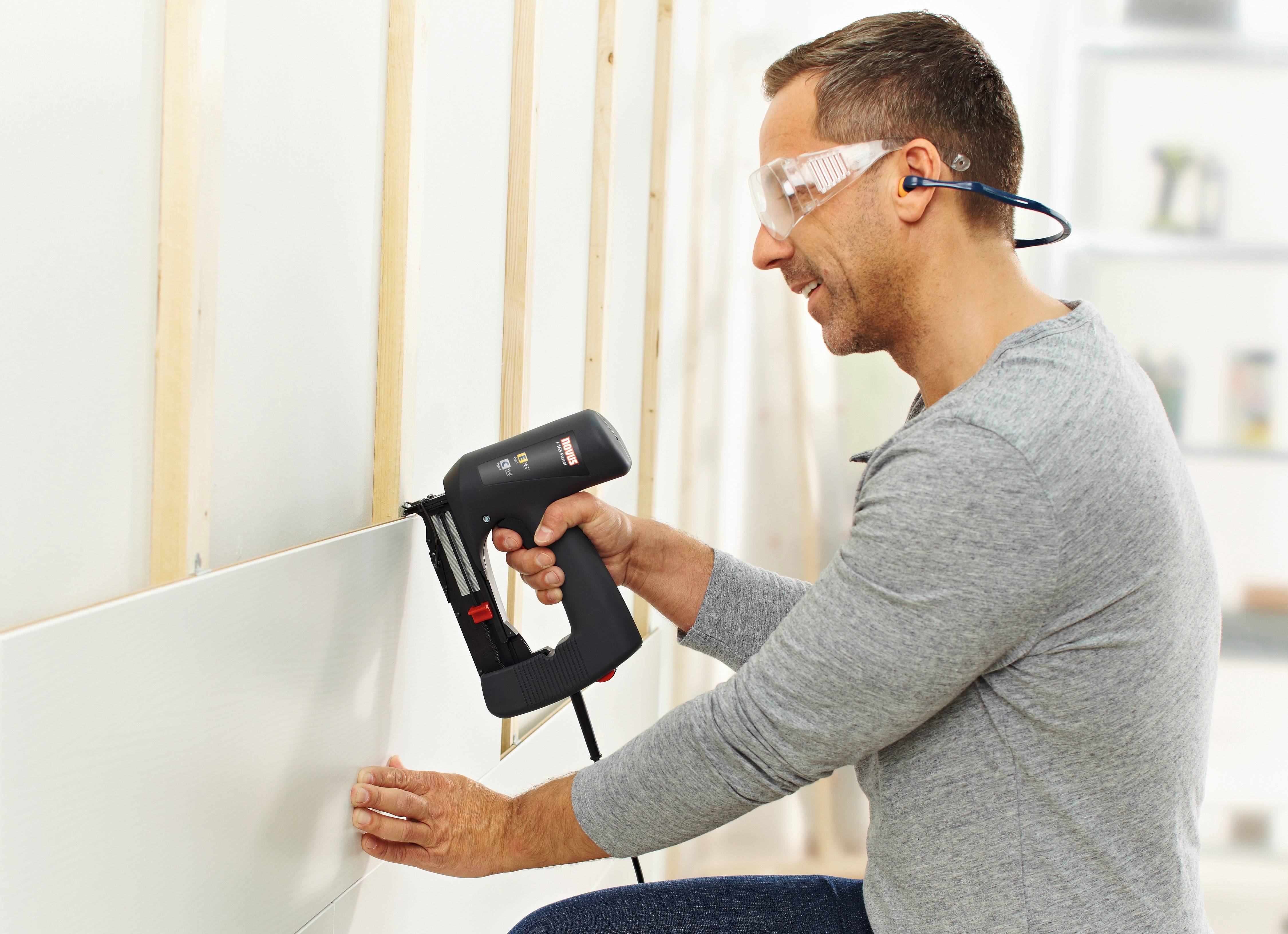 A man wearing safety glasses is fixing a wall cladding to a wooden batten using an electric staple gun in a bright room.
