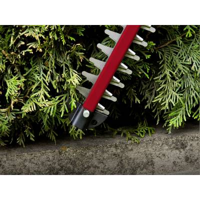 A red hedge trimmer cuts green shrubbery against a stone wall.