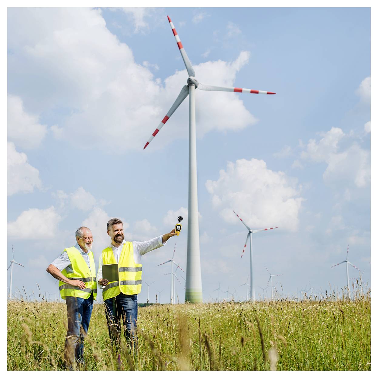 Two engineers in safety vests and helmets stand in a field with wind turbines, discussing and using a device, under a partly cloudy sky.