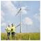 Two engineers in safety vests and helmets stand in a field with wind turbines, discussing and using a device, under a partly cloudy sky.