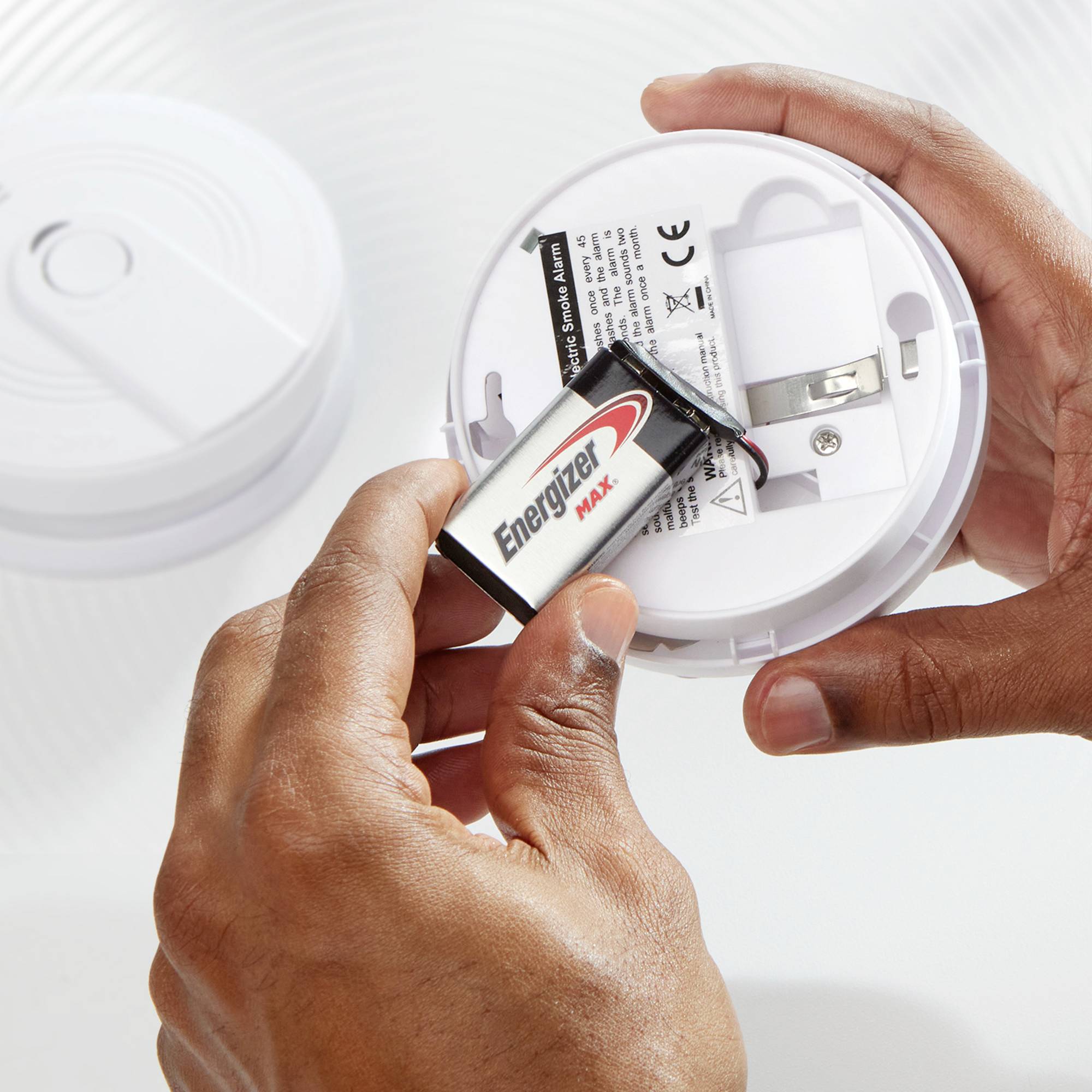A person is holding and displaying the back of a smoke detector while installing an 'Energizer Max' battery.
