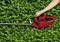A person is trimming a green hedge with a red hedge trimmer. The hedge trimmer is being held with one hand.