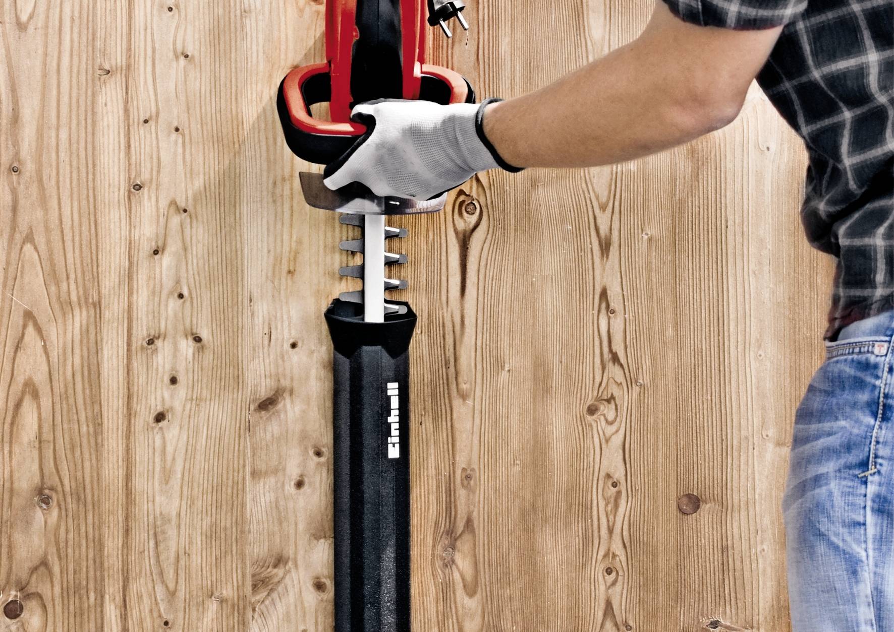 A person is using a saw to cut vertically into a wooden board. The saw is black and red, and the person is wearing gloves.
