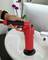 A hand is using a red plunger to clean a white sink. In the background, an orchid is placed on a shelf.