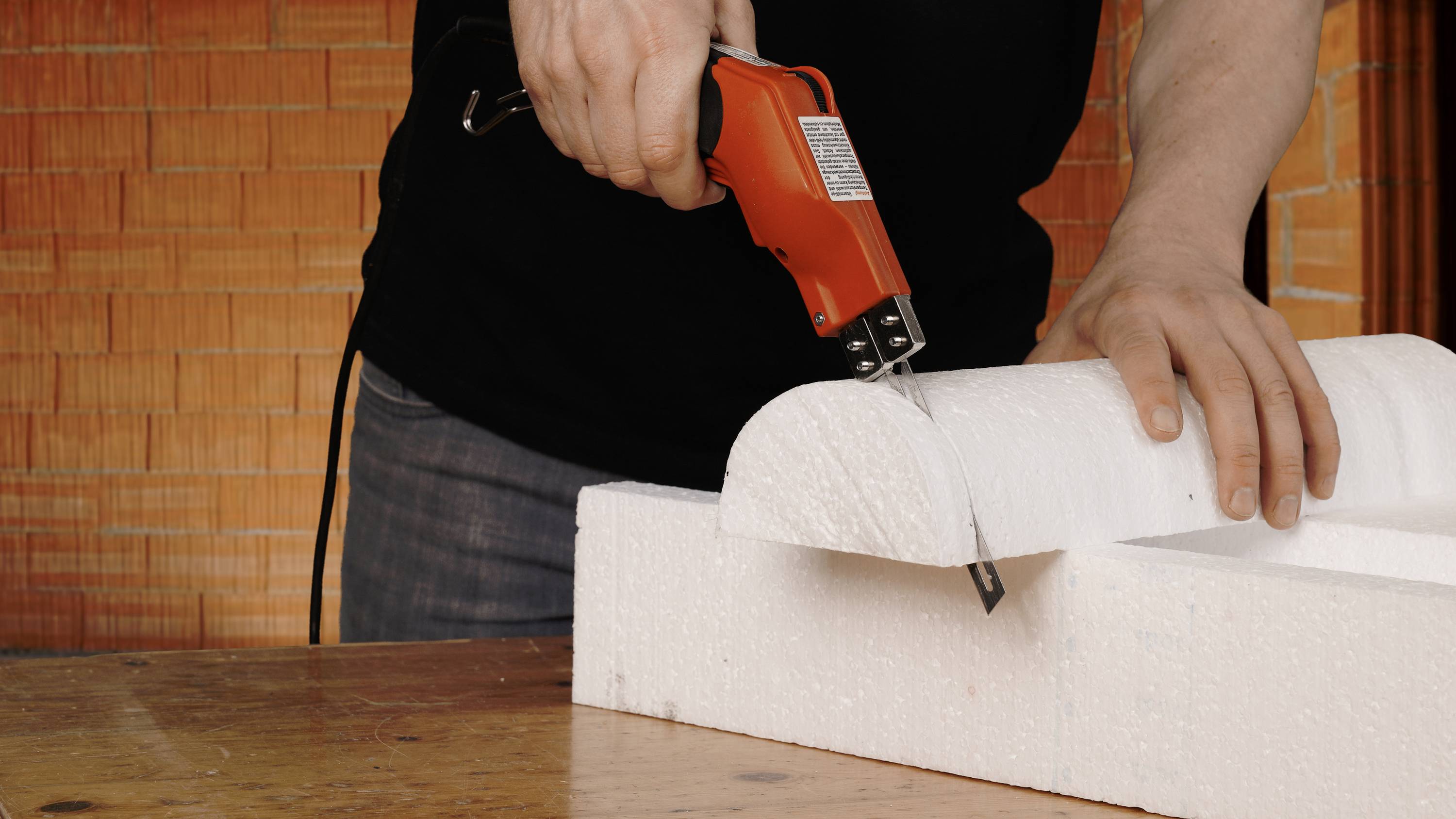 A person is cutting polystyrene with a red hot wire cutter. Brick walls are visible in the background.