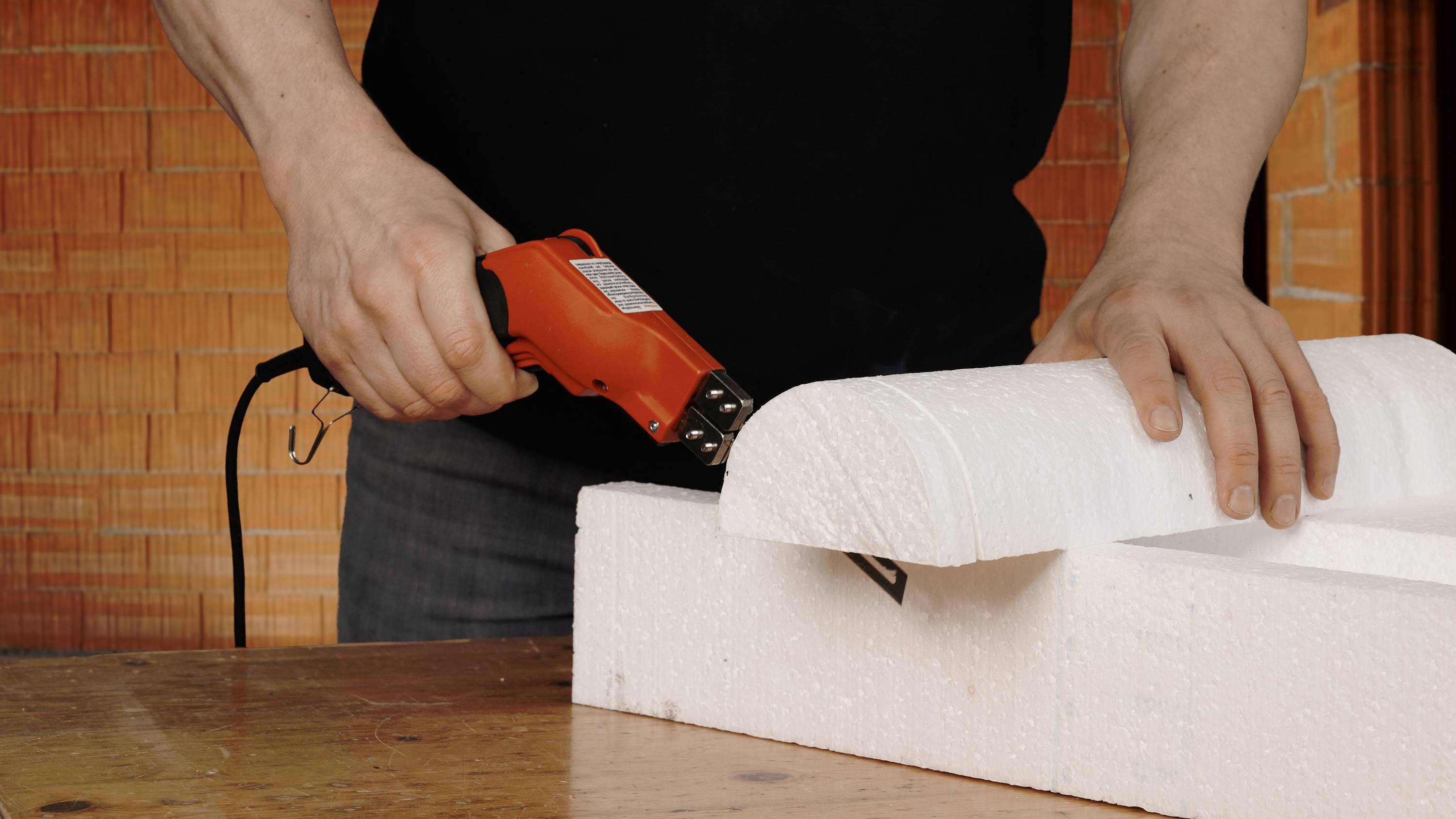 A person is cutting polystyrene with an electric cutting tool on a wooden table.<br><br>Note: In British English, "Styropor" is typically referred to as "polystyrene", and the phrasing is kept natural and concise.