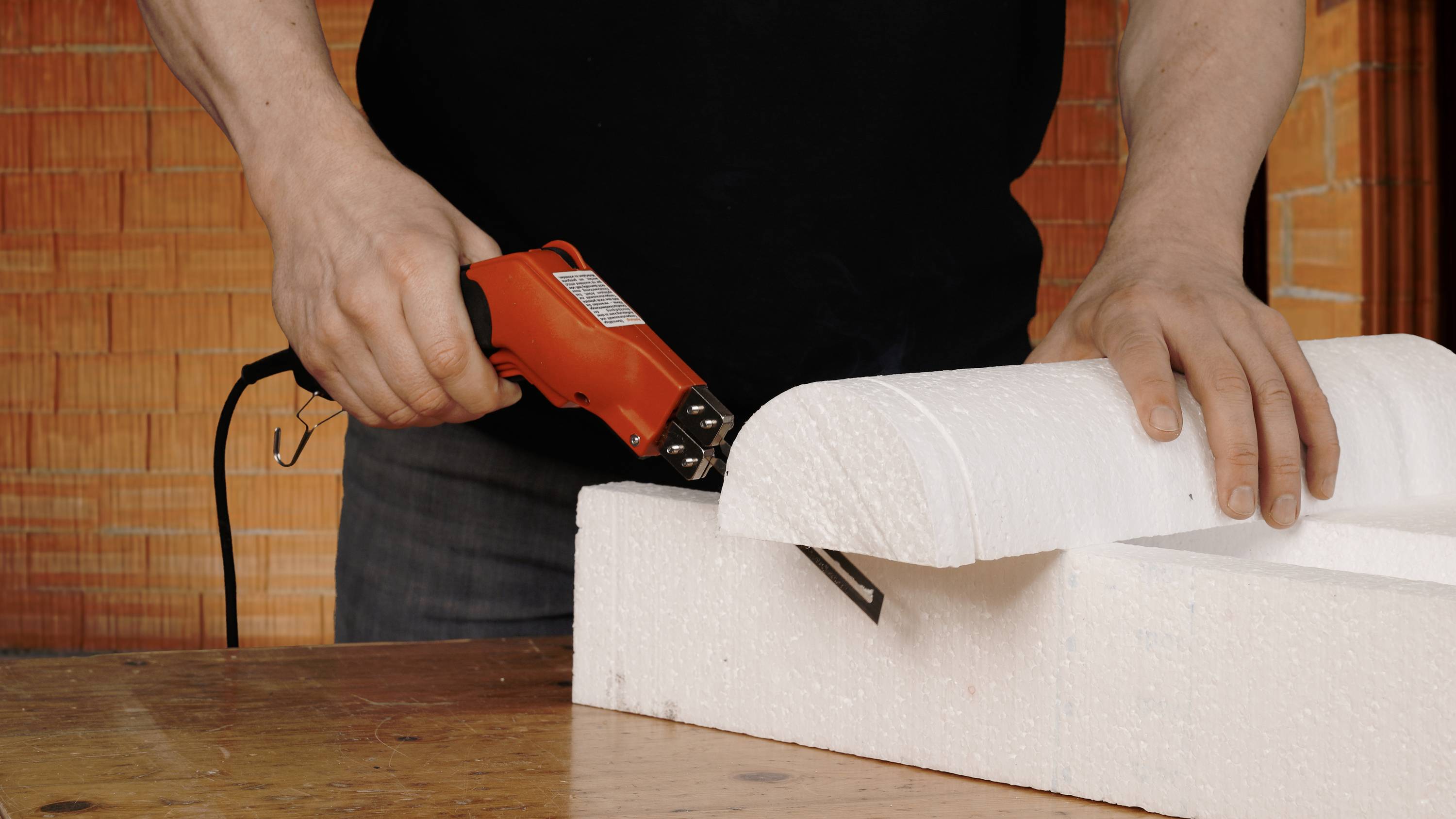 A person is precisely cutting through a thick piece of white polystyrene with an electric cutting tool on a wooden table.
