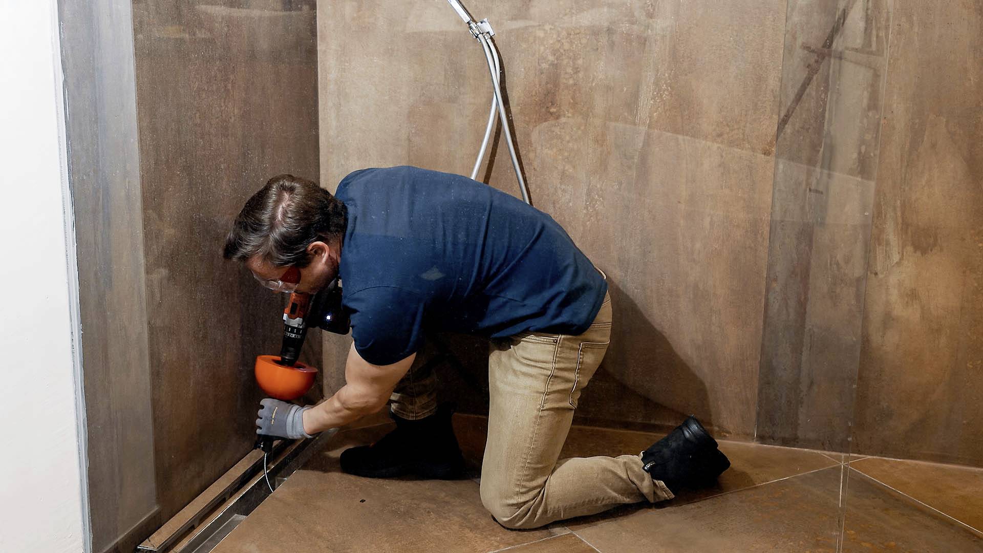 A tradesman is installing a shower drainage channel in the bathroom. He is kneeling on the floor and working on the drainage channel with a tool.