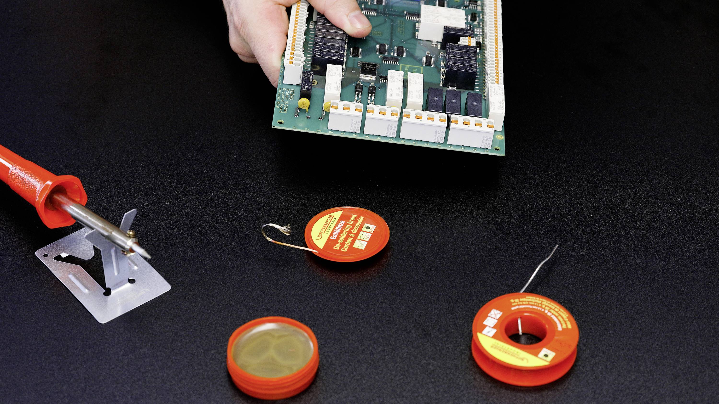 A person is holding an electronic circuit board over a black table equipped with a soldering iron and soldering materials.