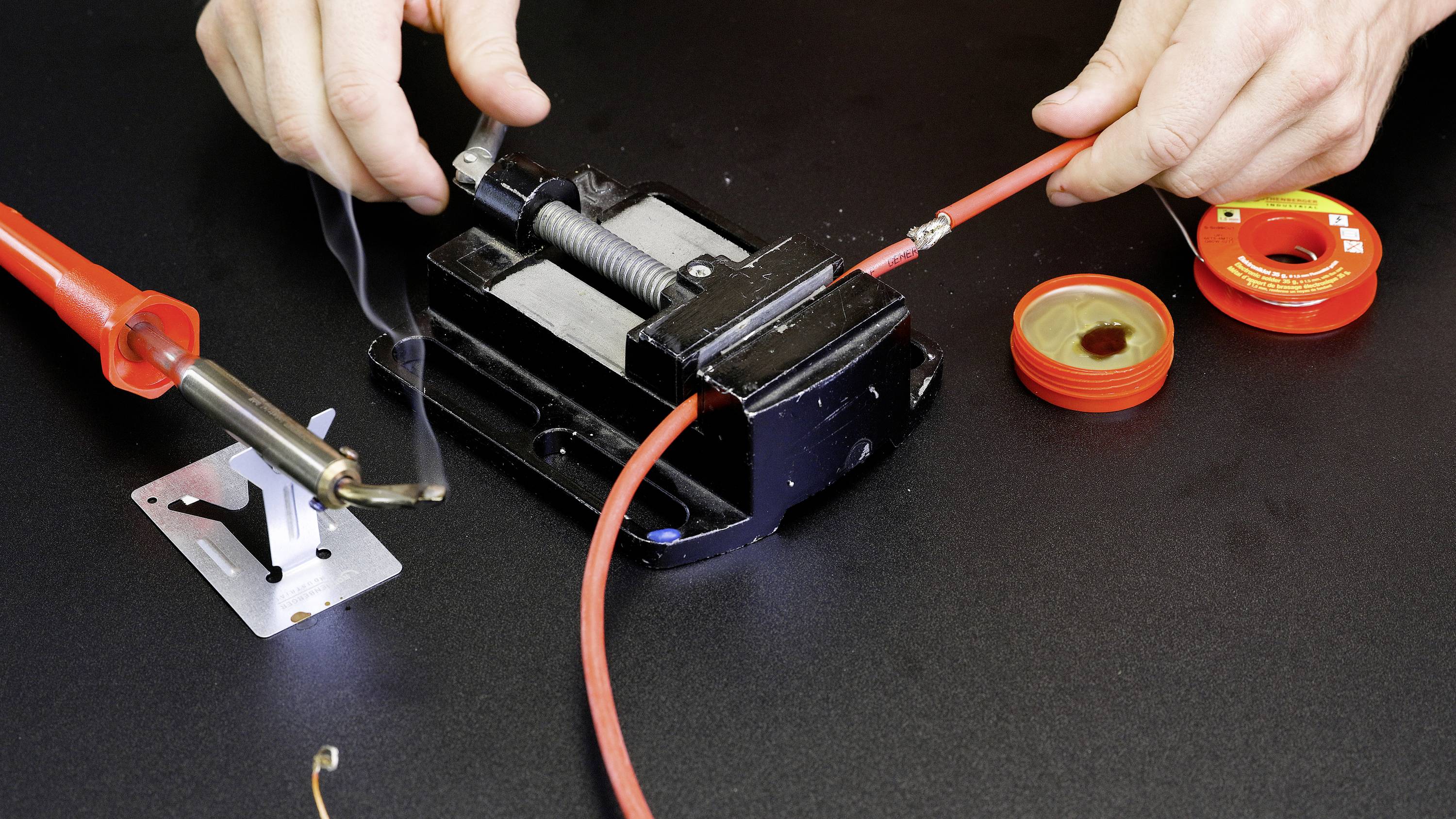 A person is soldering a red cable with a soldering iron on a black surface; smoke rises from the cable, with tools in the background.
