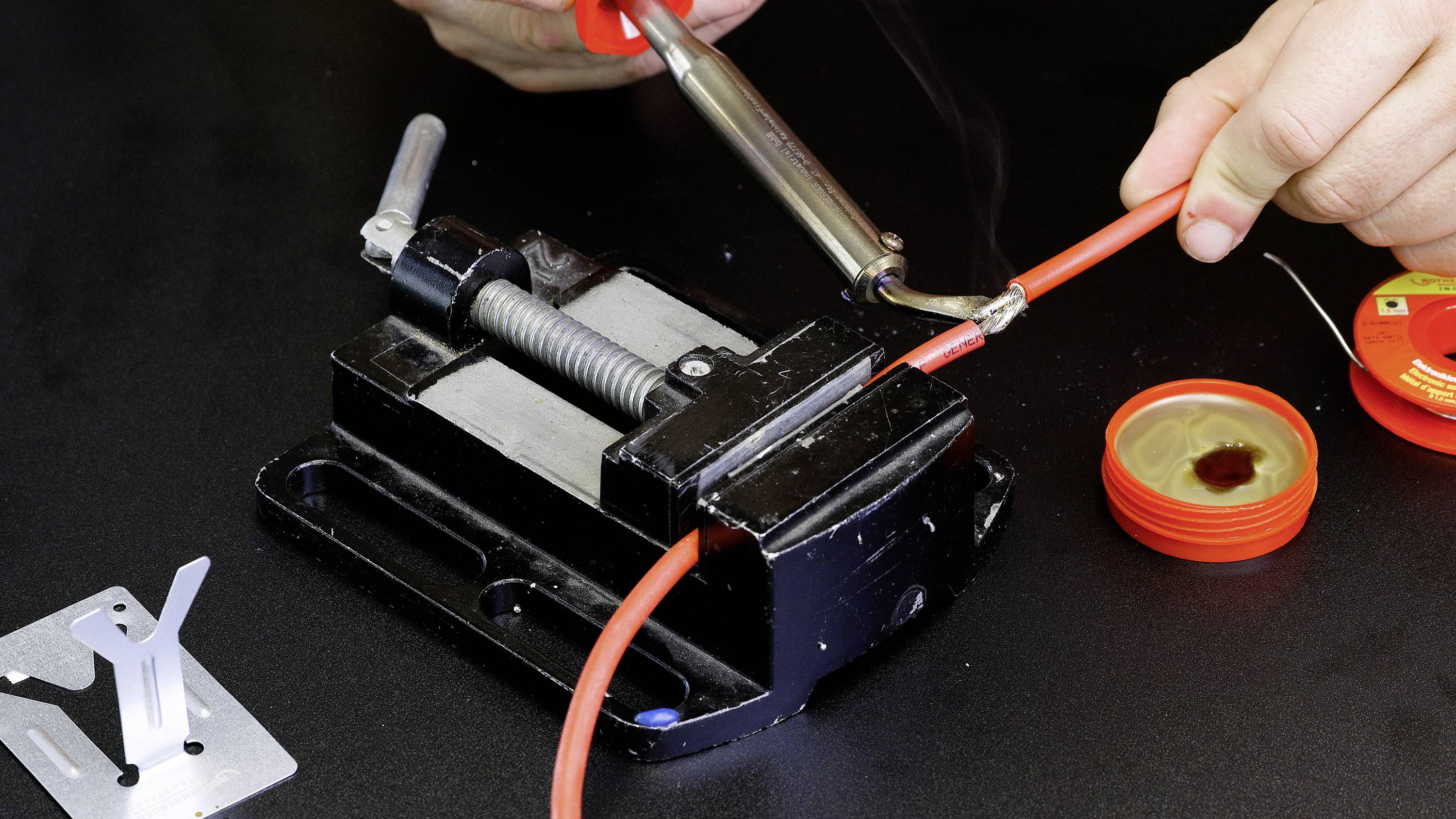 A person is soldering a red cable that is clamped in a vice. Solder wire and an open flux container are lying nearby.