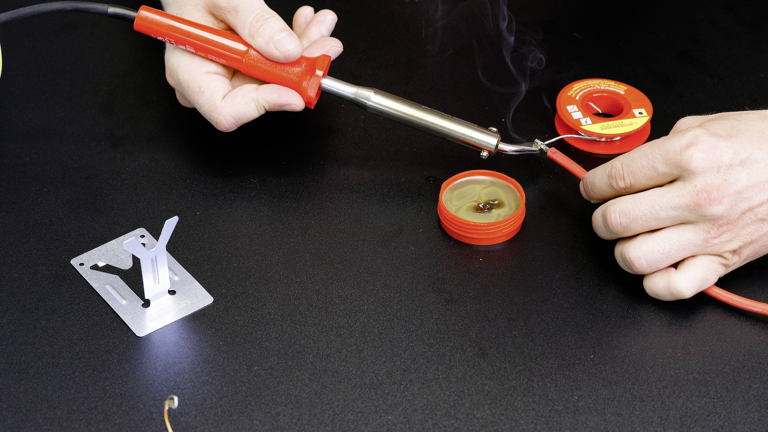 A person is soldering a cable with a soldering iron. Next to the cable, solder and soldering accessories are lying on a black table.