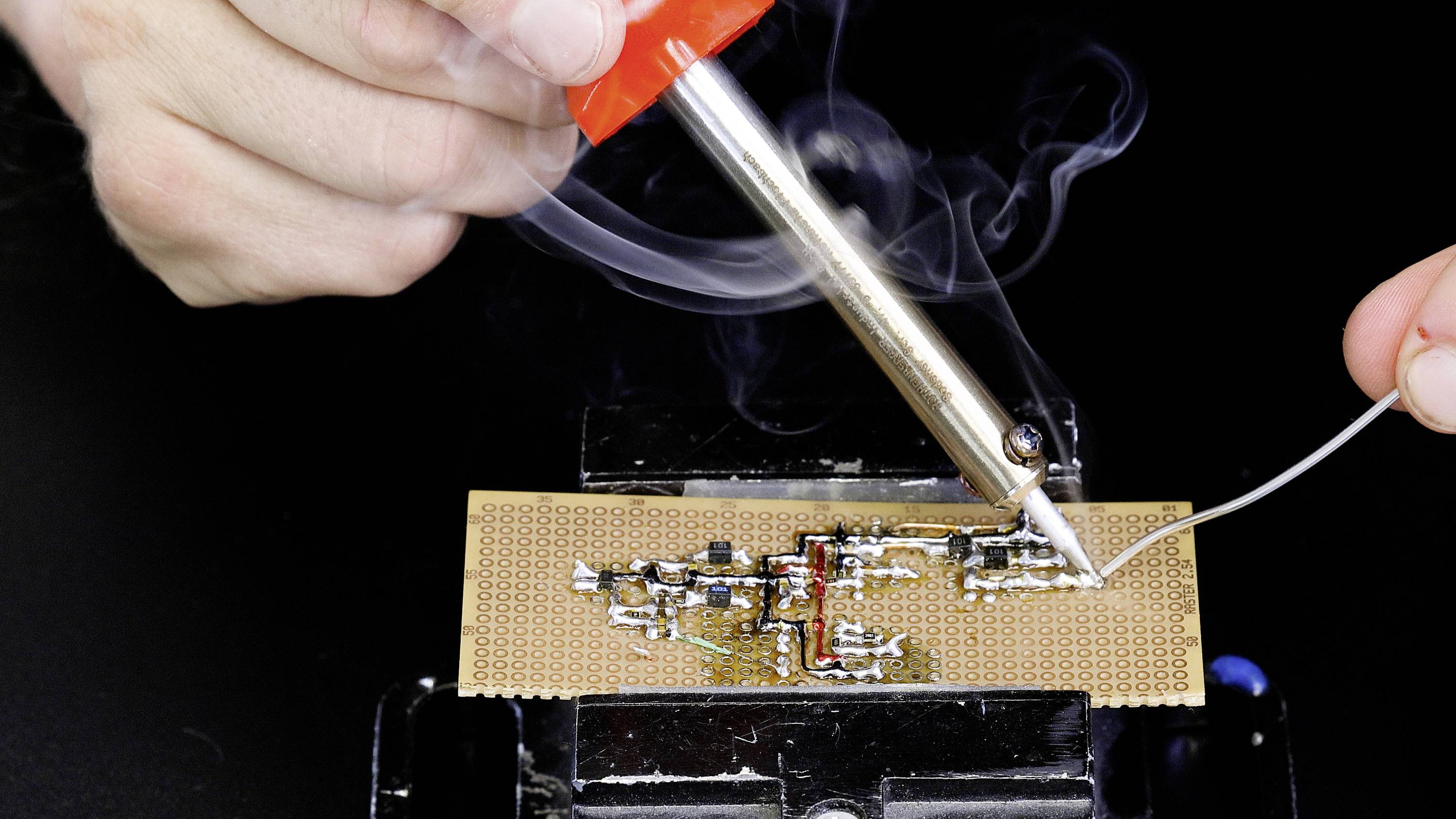 A hand solders electronic components onto a circuit board. Smoke rises from the hot soldering iron while the board is secured in place.