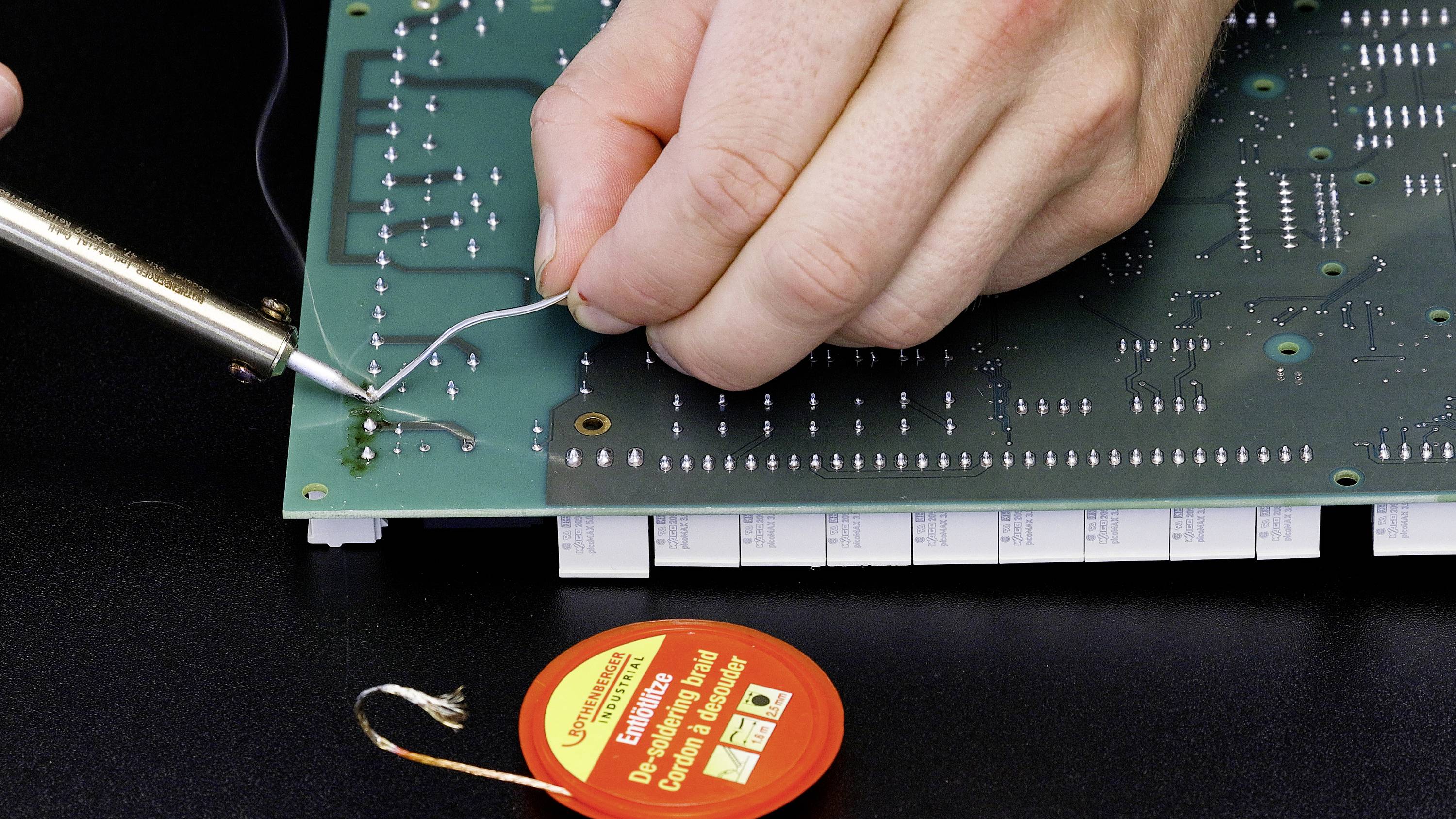 A person is soldering a circuit board with a soldering iron and solder. A tin of flux is lying next to it on the work surface.
