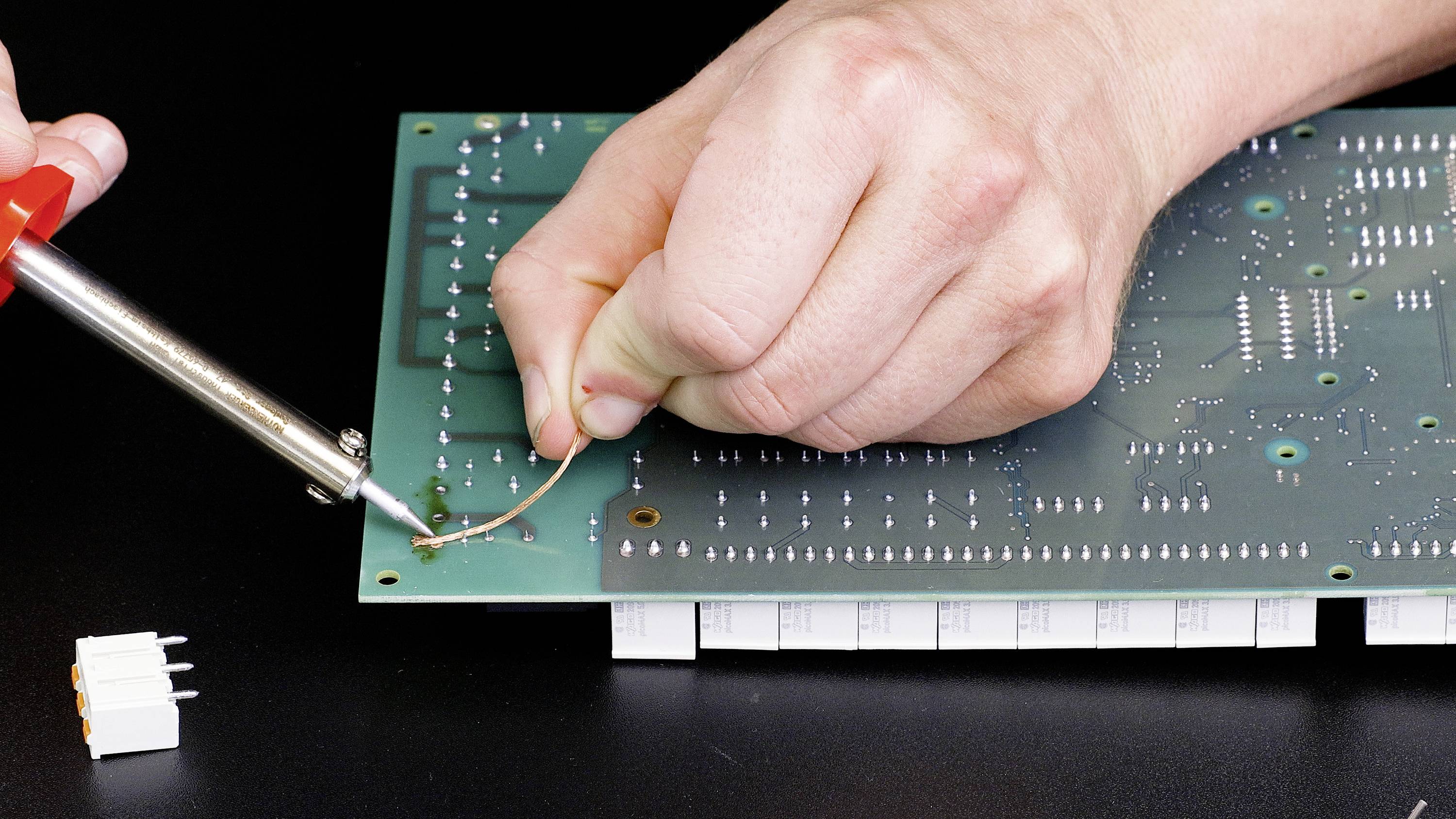 A person is soldering a circuit board with a soldering iron. A small connector lies beside it.