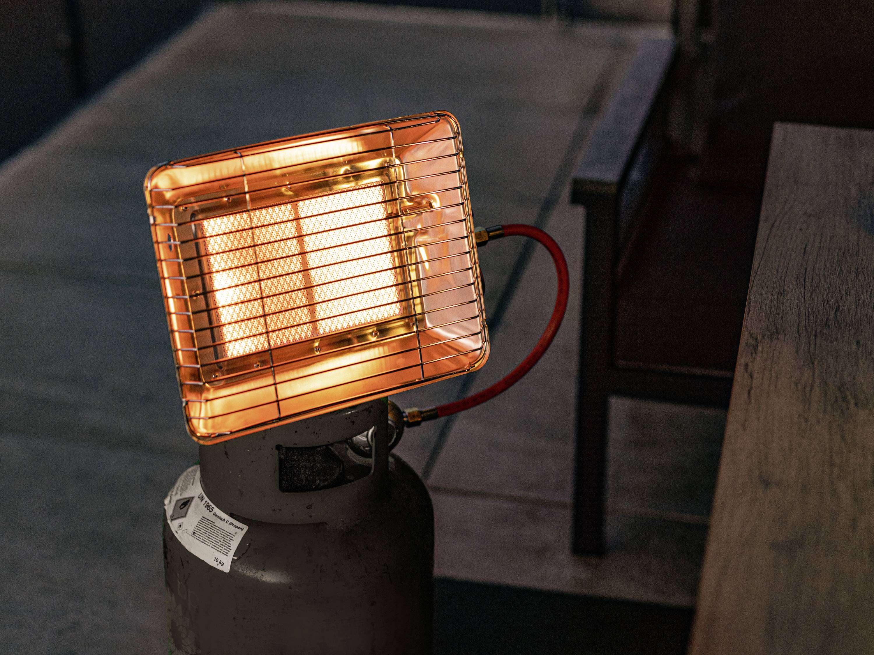 An infrared heater on a gas cylinder stands on a floor beside a wooden table and glows orange in a dark room.