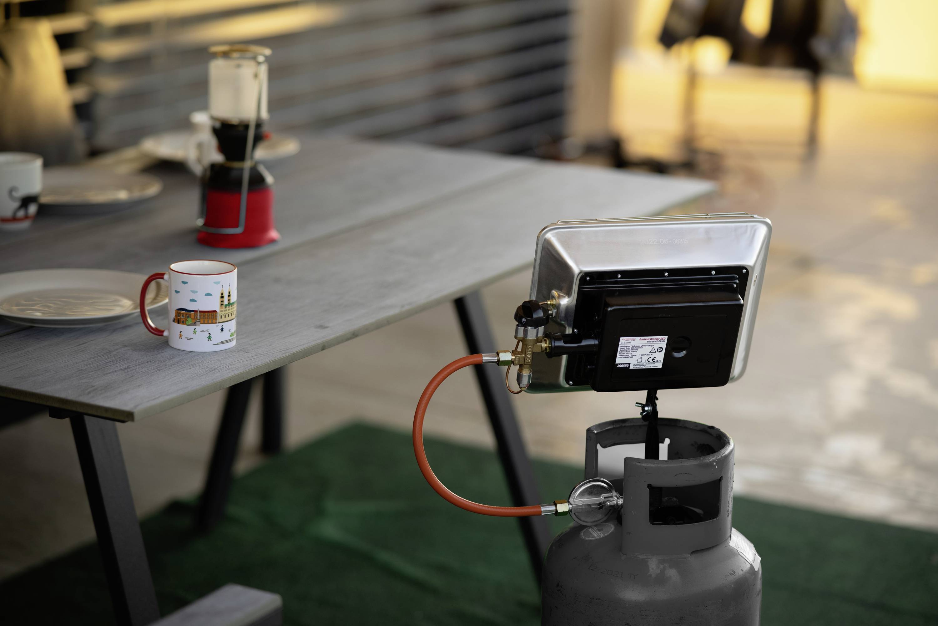 A gas heater on an outdoor table, connected to a gas bottle. A cup and other pieces of crockery are sitting on the table.