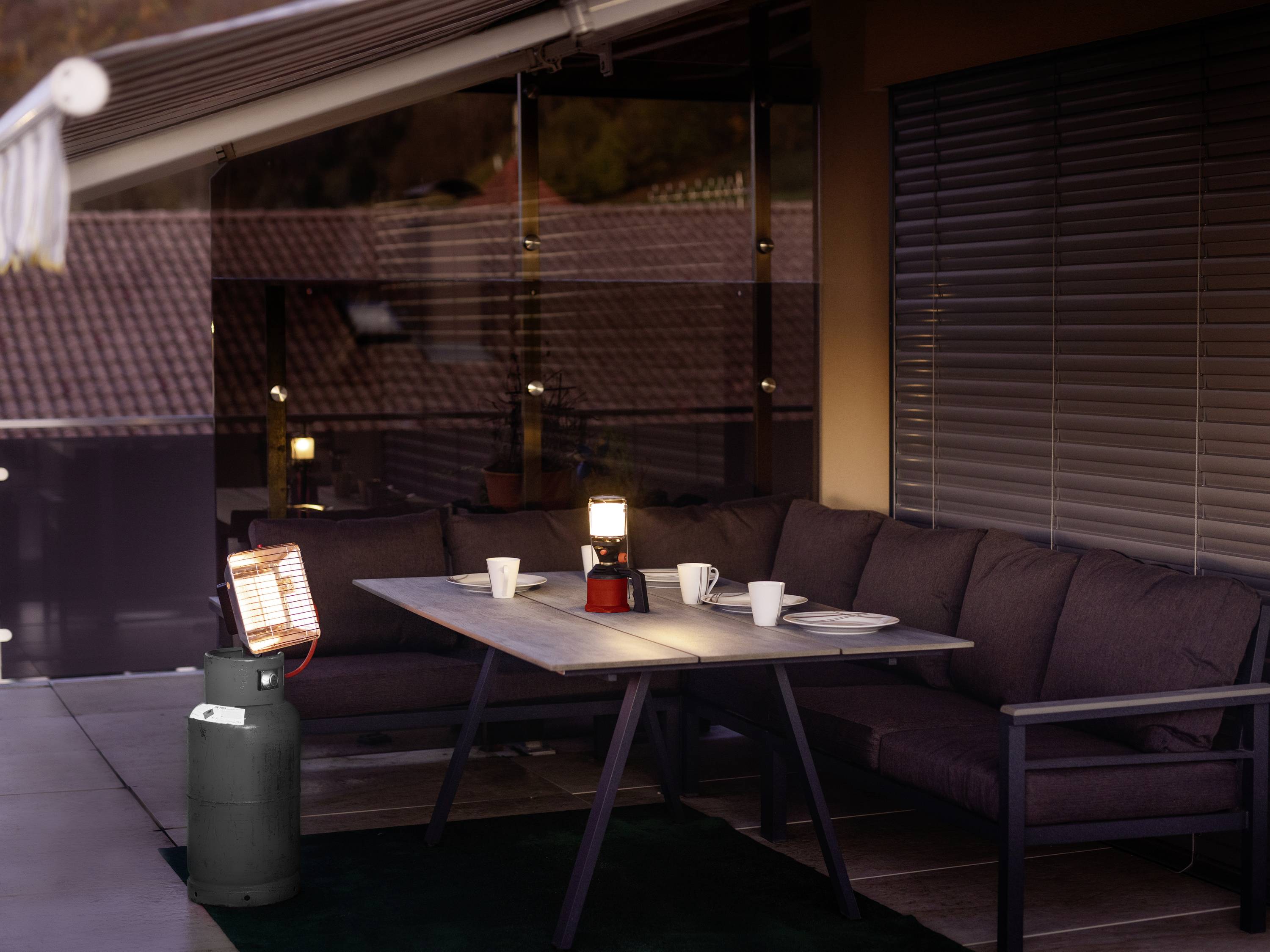 A terrace at dusk with a sofa and table. A gas heater, a lantern, and mugs are placed on it. Windows reflect the surroundings.