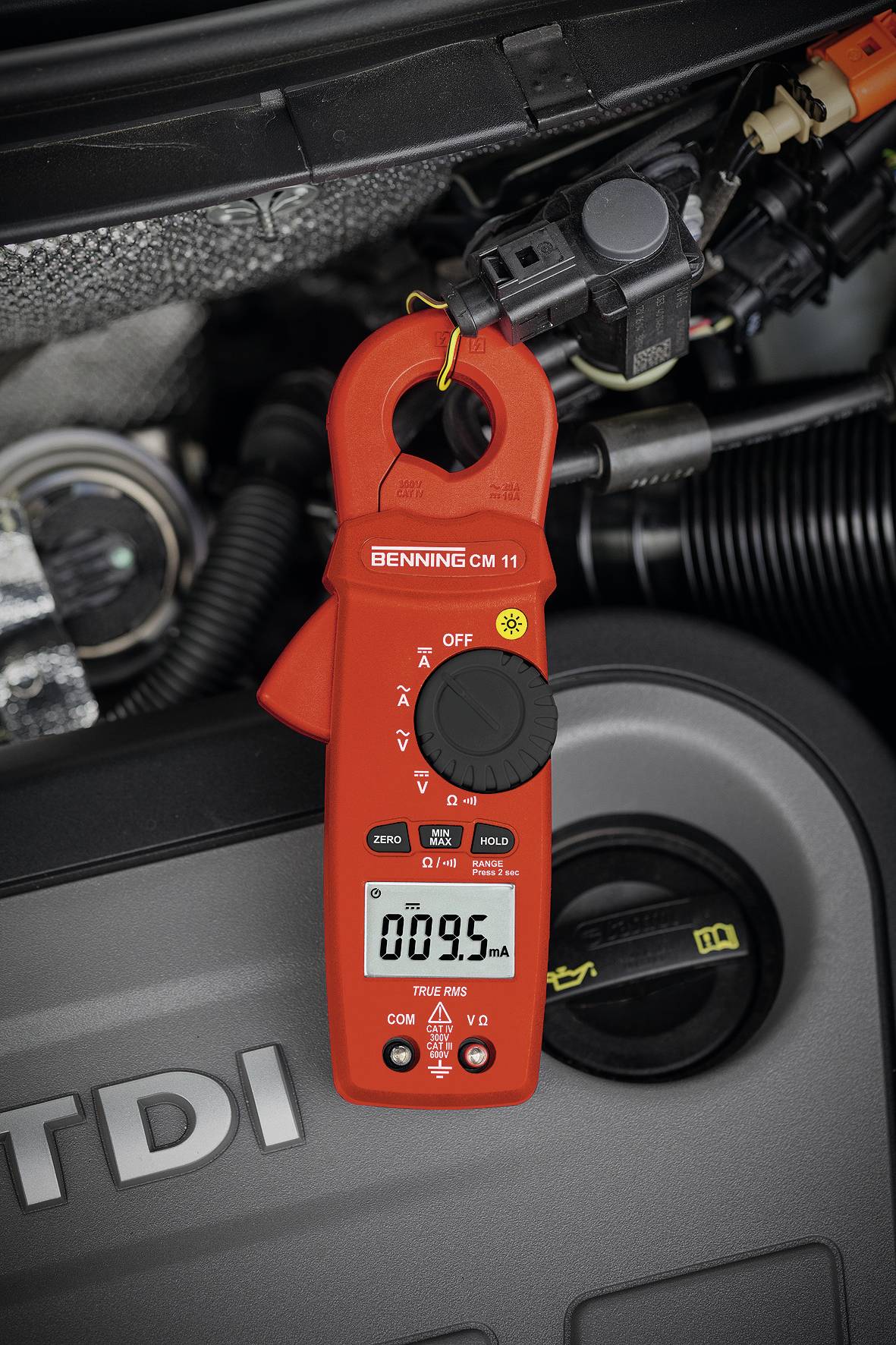 A red multimeter displays 009.5 mA and is attached to a cable in a car workshop. Engine components are visible in the background.