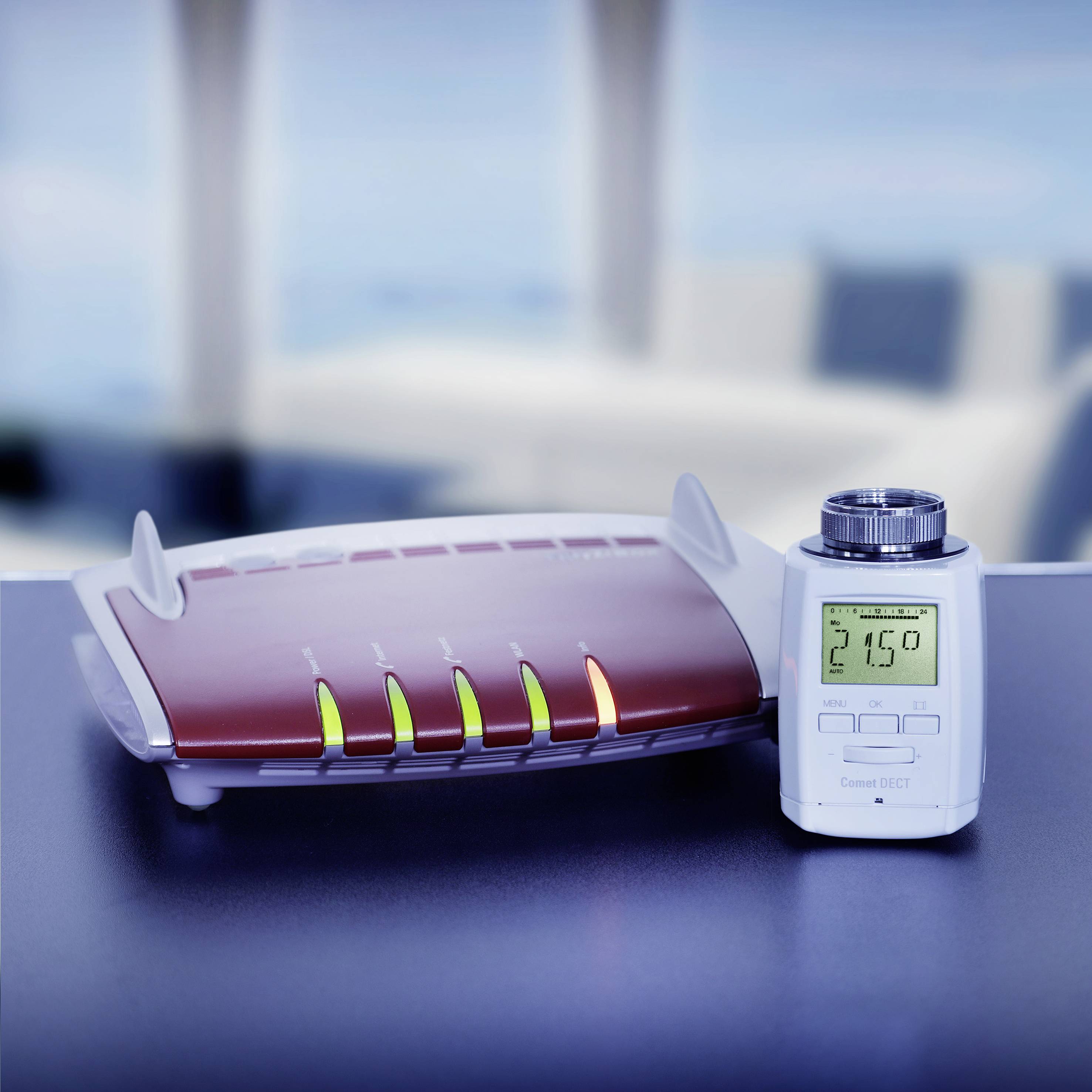 A wireless router sits on a table, next to a small digital thermometer displaying 21.5 degrees Celsius. A blurred room can be seen in the background.