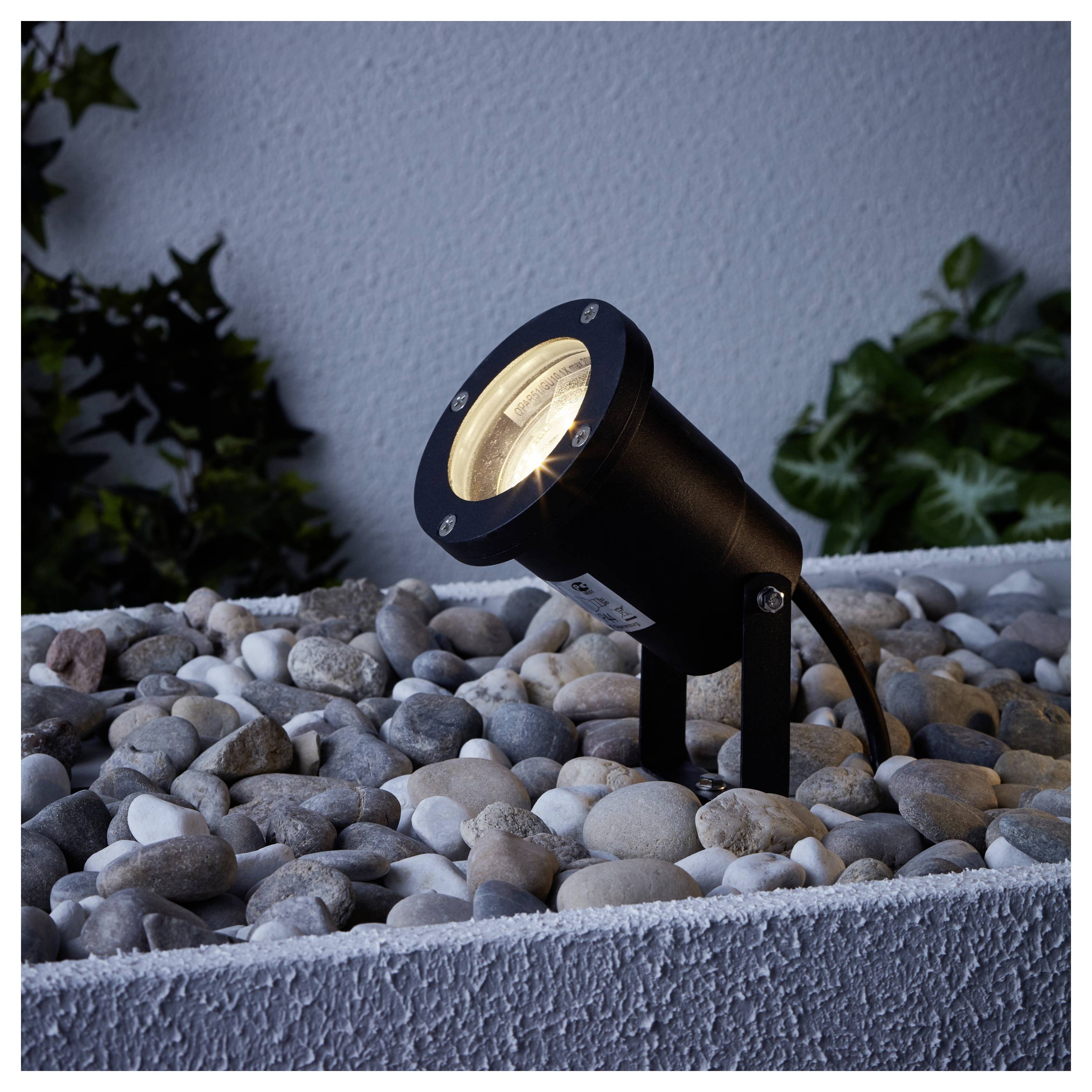 A black outdoor spotlight illuminates a bed of smooth pebbles against a white wall, highlighting nearby green foliage.