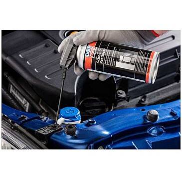 A person's gloved hand sprays a can of lubricating spray onto a car engine's blue metal component, aiming to prevent squeaks.