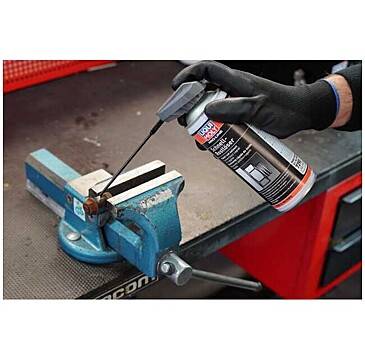 'A hand wearing a glove sprays lubricant on a metal part held in a blue clamp on a workbench.'