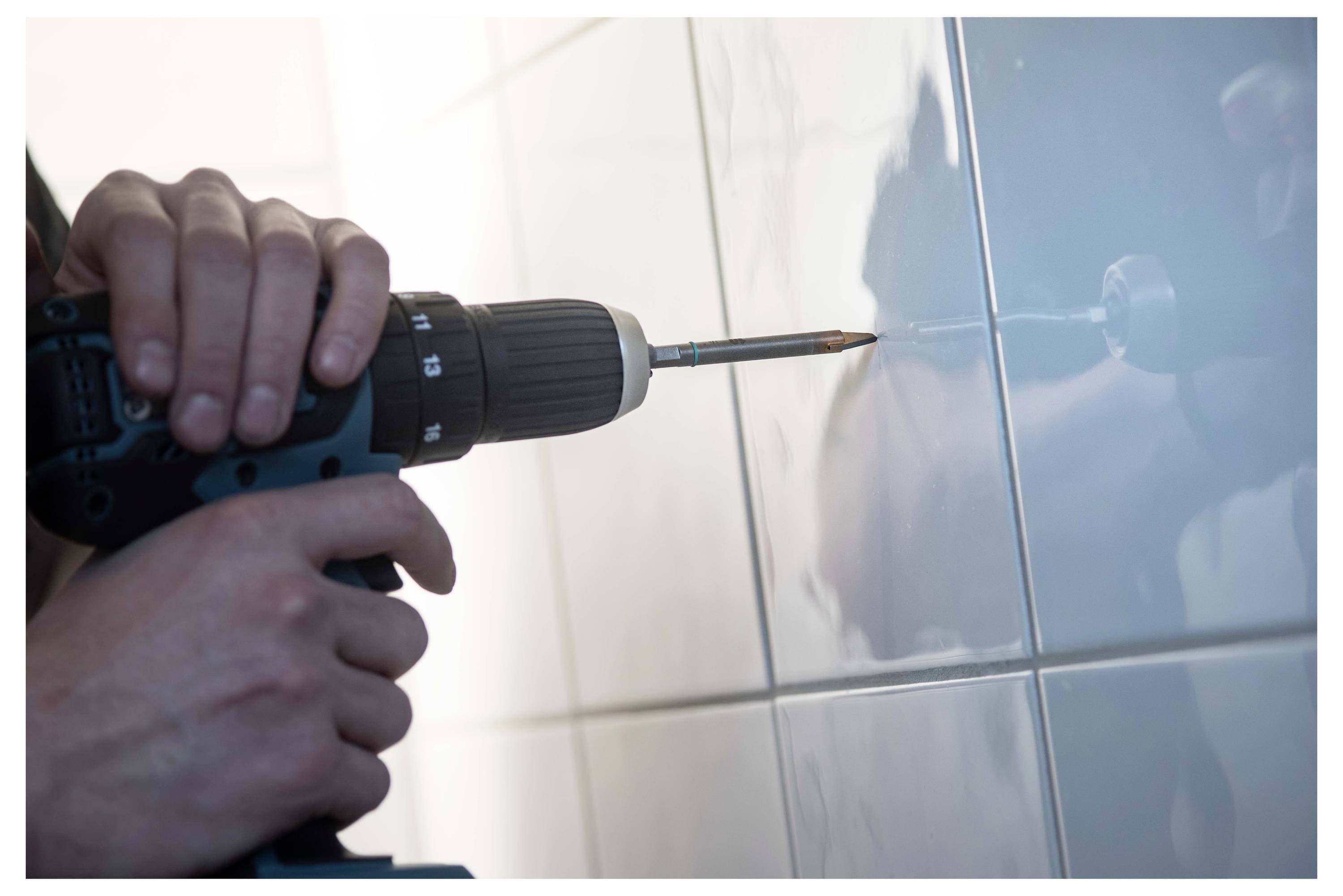 Hands holding a cordless drill with a tile bit, drilling into a glossy tiled wall.