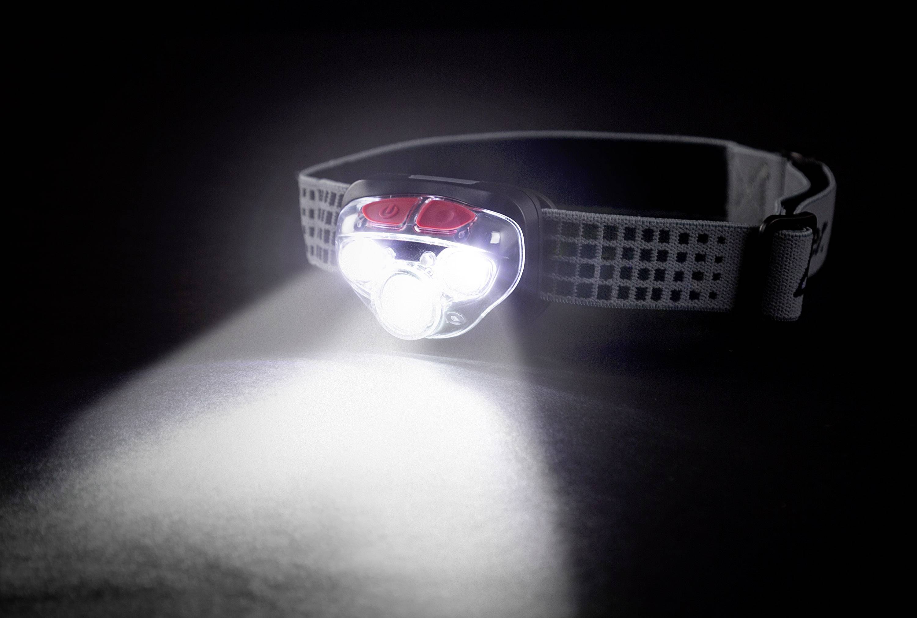 A head torch illuminates a dark room, with the beam of light casting forward. The strap is adjustable and grey with a pattern.