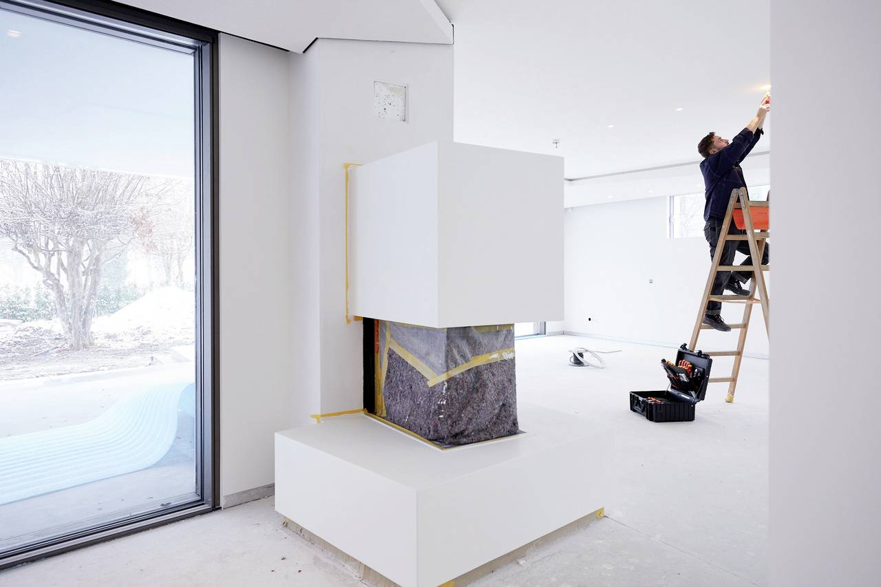 A person on a ladder is installing lighting in a modern interior. A large white cube is in the room, near a window.