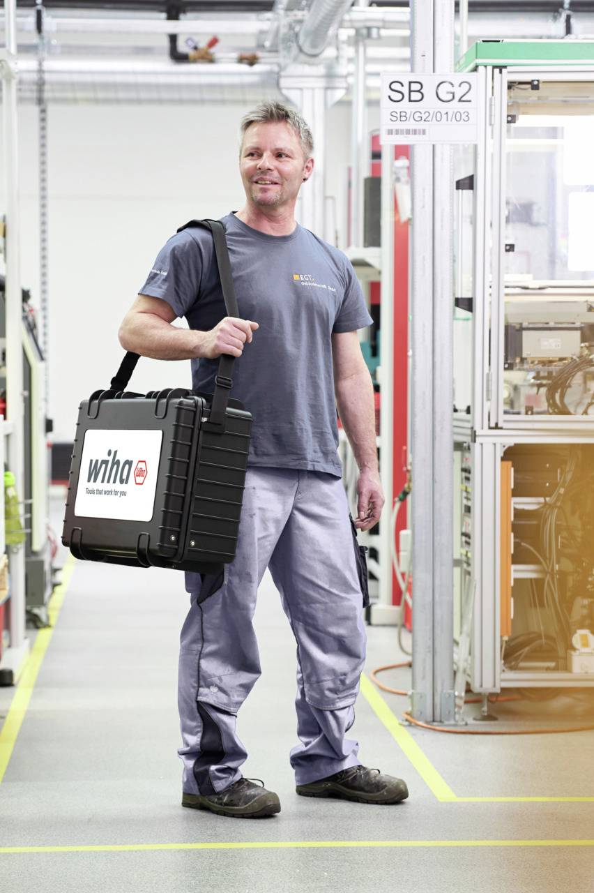 A man is standing in a workshop, carrying a black tool case with a 'wiha' logo. He is wearing a grey T-shirt and work trousers.