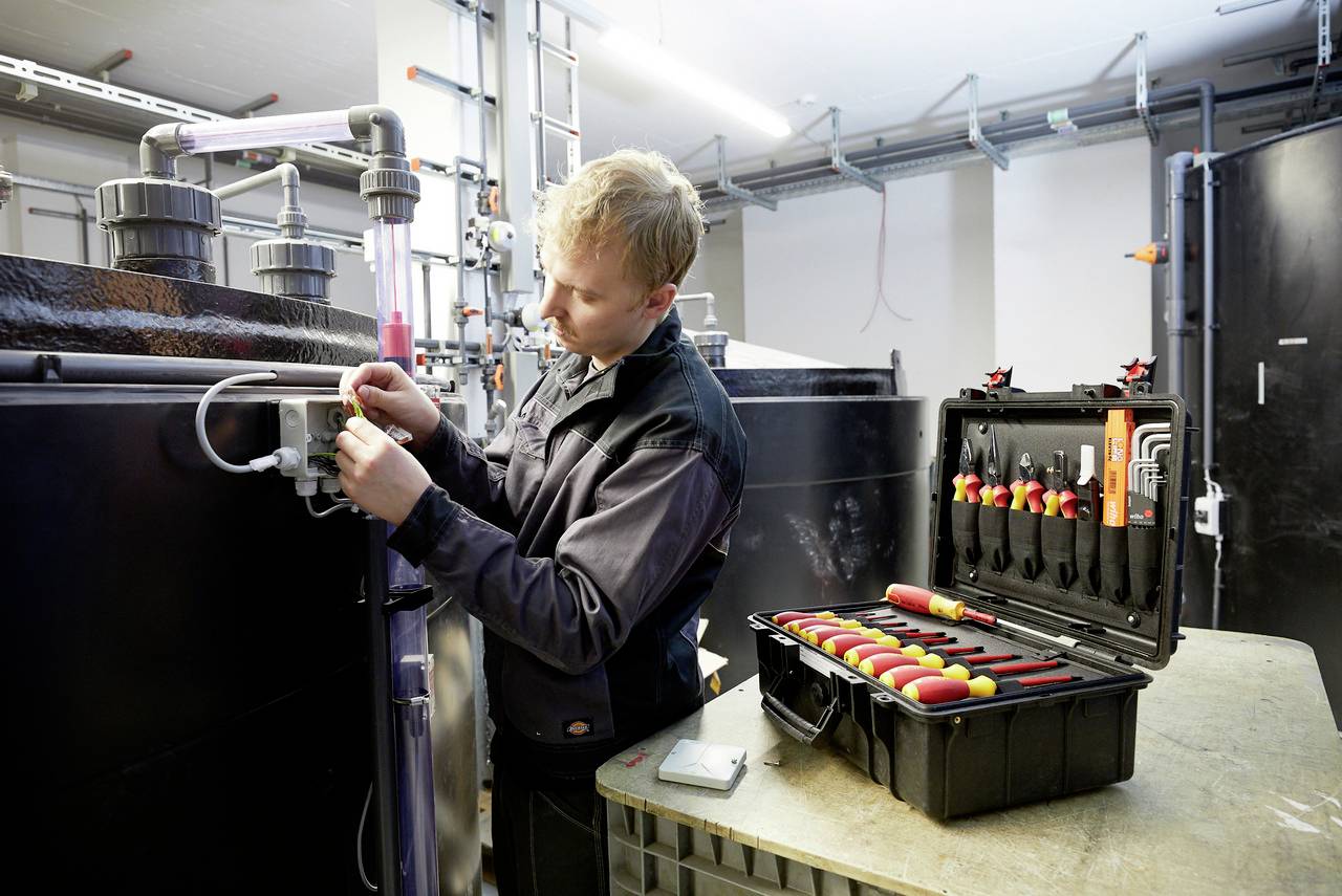 A technician is working on a large tank, using tools from an open toolbox. Pipes and pipework are visible in the background.