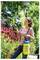 A person uses a garden sprayer to water red flowers in a sunny park. They hold a yellow and blue sprayer with 'Gloria' branding.