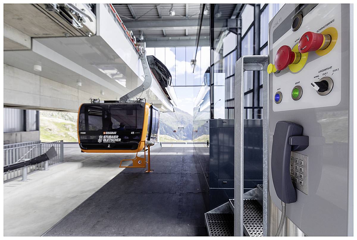 Cable car station with gondola; control panel with emergency button in the foreground; large windows offer a view of alpine landscape in the background.