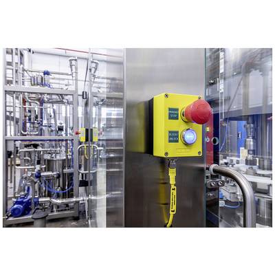 An industrial facility with an emergency stop switch in the foreground. The switch is yellow with a red button, surrounded by machinery.