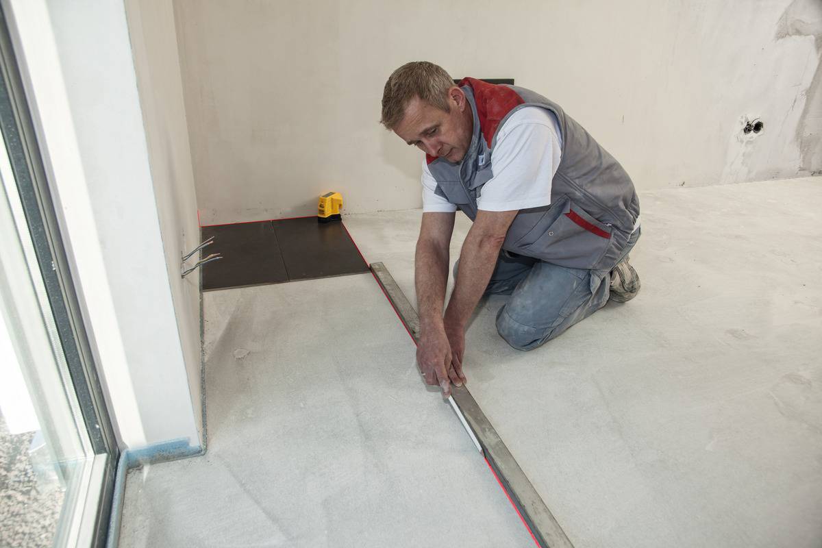 A person is kneeling on a floor and measuring with a tape measure. A spirit level and cables are visible in the background.