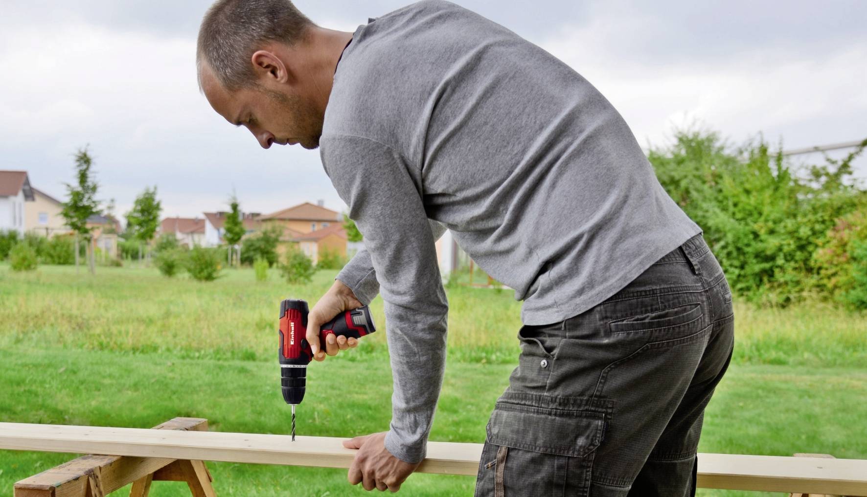 A man is using an electric drill in a garden to drill holes into a wooden board. Houses and trees can be seen in the background.