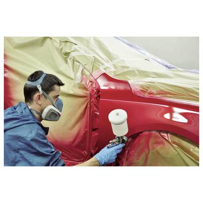 Technician spray-paints a red car panel in a body shop, using a spray gun while the rest of the vehicle is masked with protective plastic and tape.