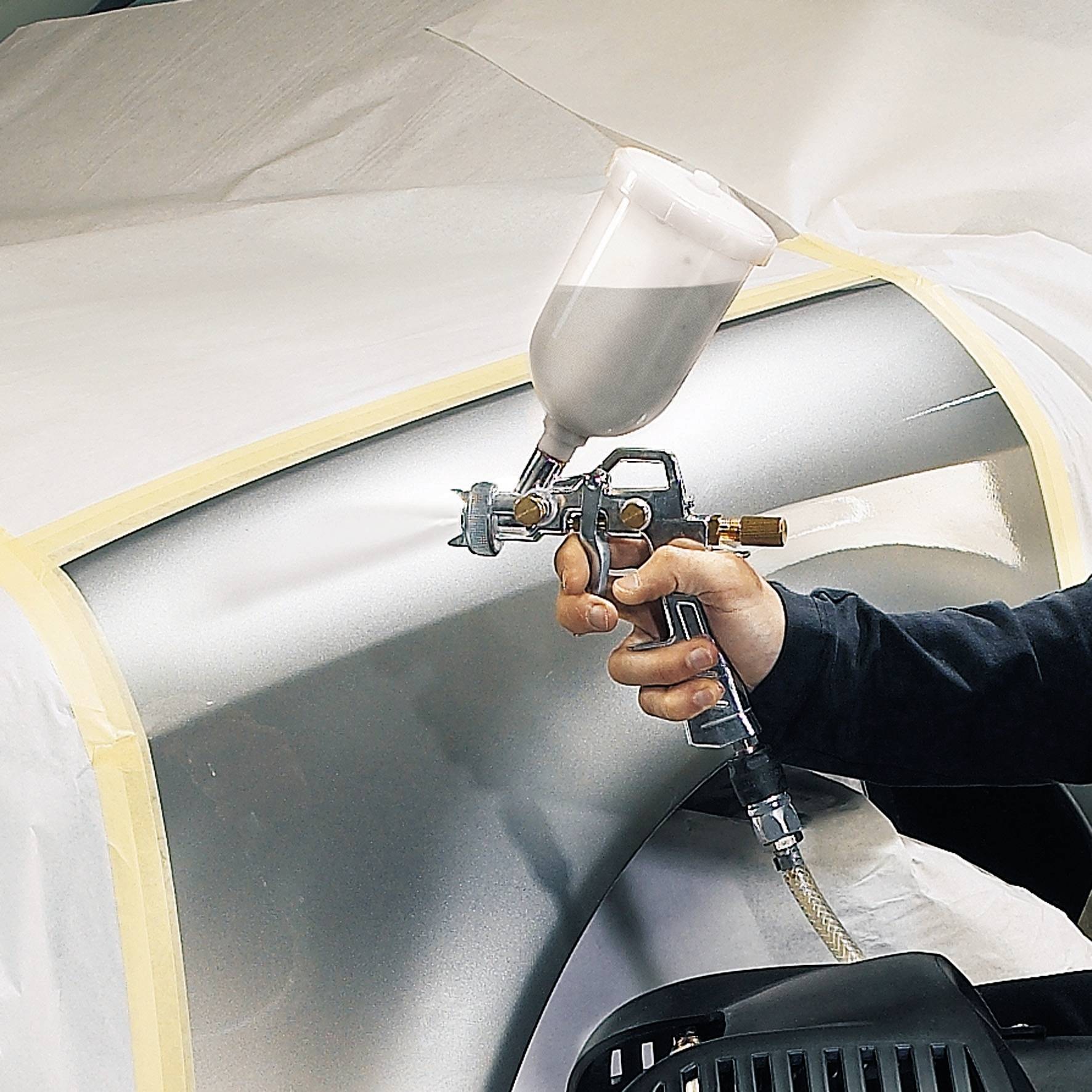 A hand is spraying white paint with a spray gun onto a masked car part. The image depicts a paintwork task.