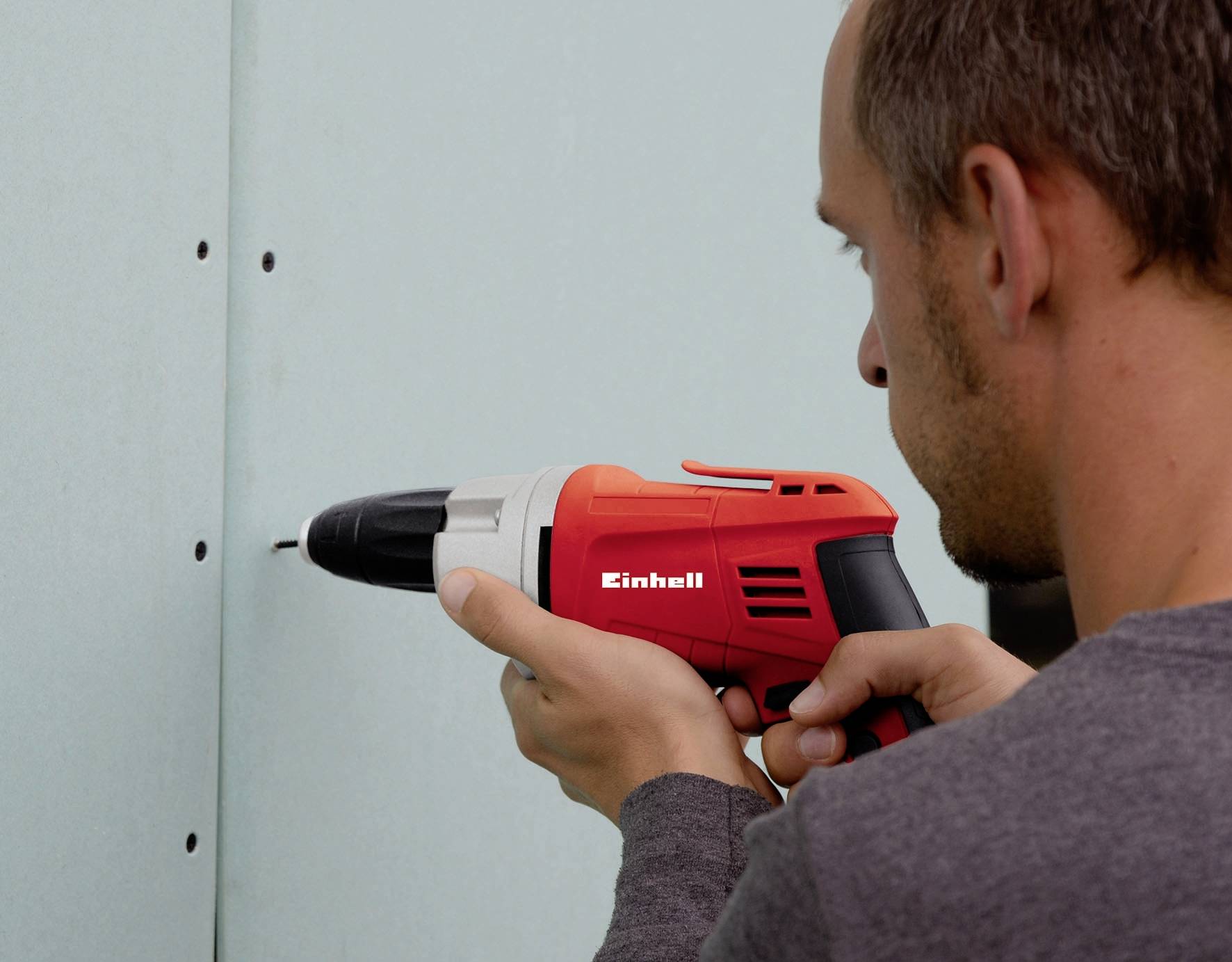 A man is installing a green plasterboard sheet to a wall using a red and white cordless drill.