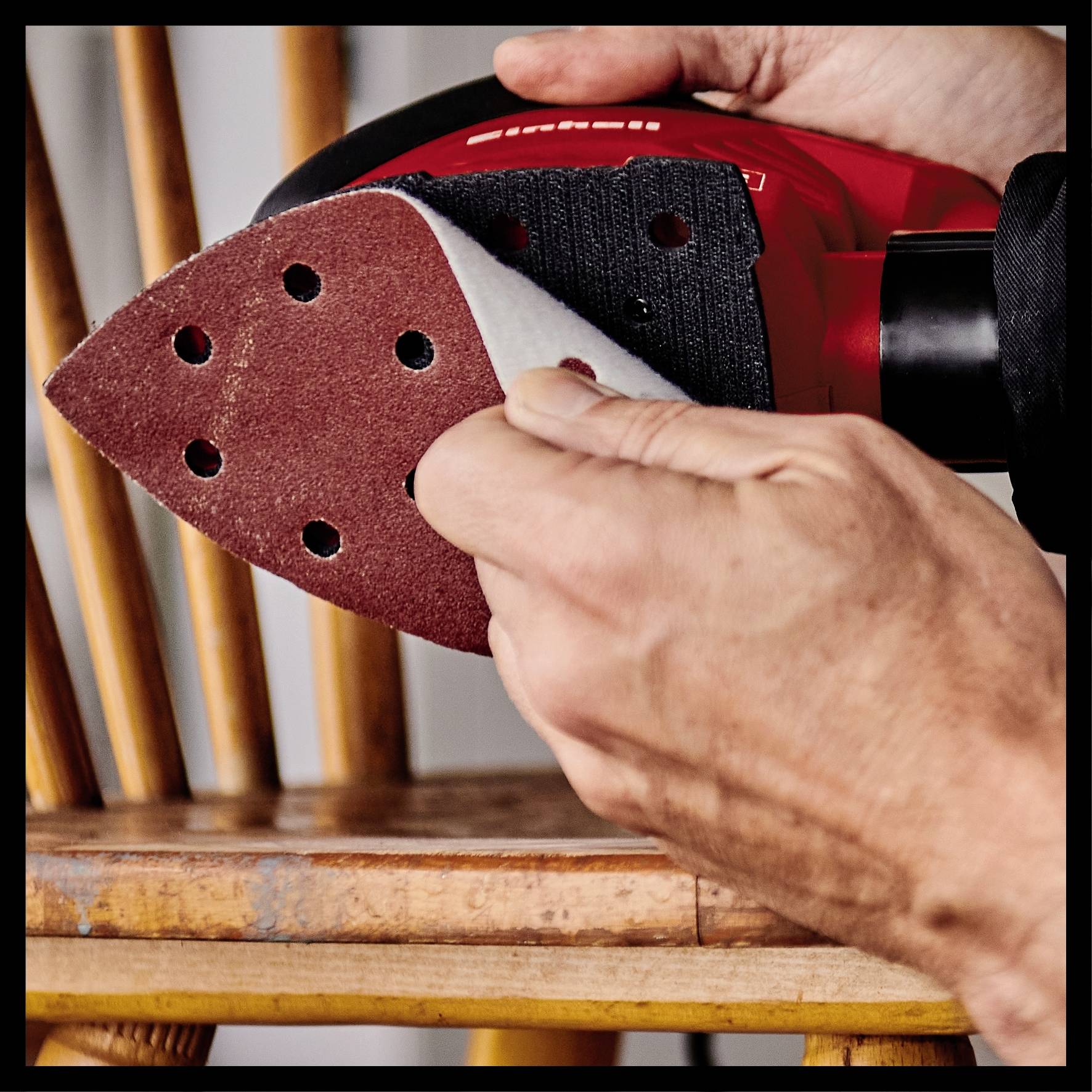 'Close-up of a hand sanding, using sandpaper on a chair. One hand holds a red orbital sander with sandpaper attached.'